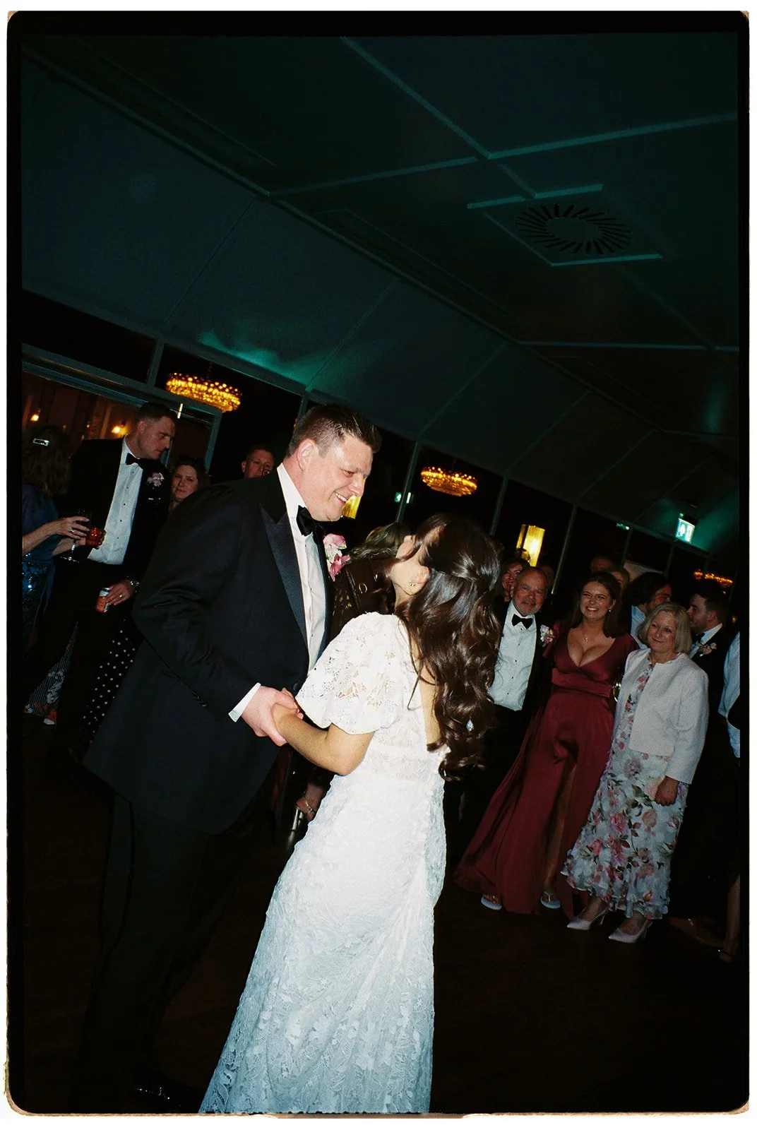 A bride and groom are dancing together at a wedding reception, surrounded by friends and family in formal attire.