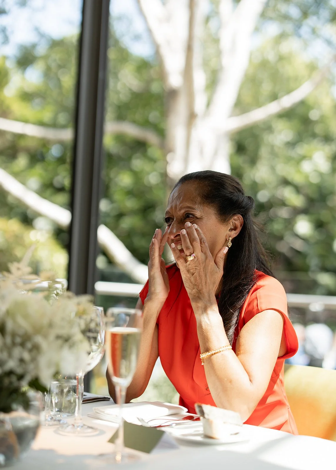 A woman in a red dress is sitting at a table with a white tablecloth, covering her face with her hands, at a celebration or special occasion, with trees visible outside through large windows.