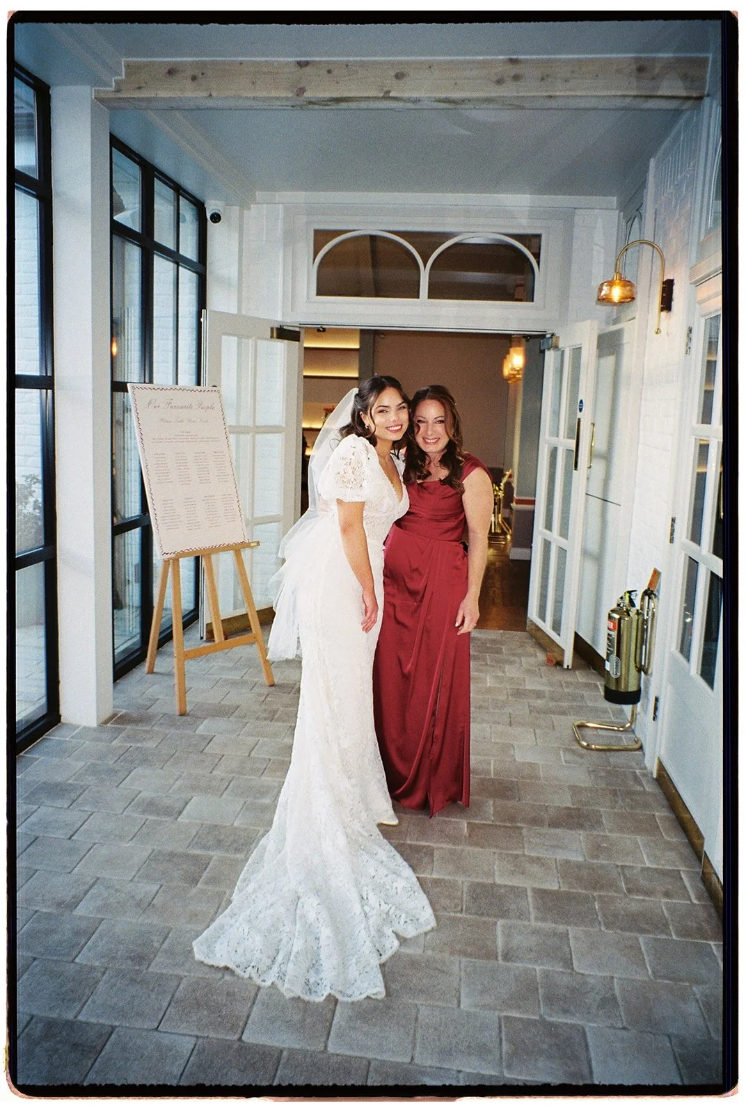 Two women, one in a wedding dress and the other in a red gown, smiling and posing inside a bright, modern venue.