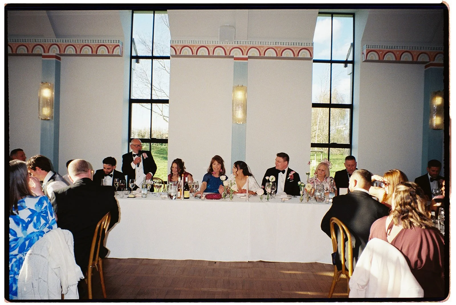 Wedding reception with a bride and groom sitting at a head table, a man giving a speech, and guests seated around in a bright room with large windows.