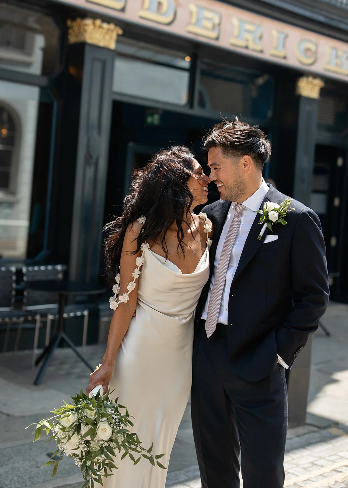 A bride and groom smiling and touching foreheads outside a building with the words "BEERICK" partially visible, celebrating their wedding day.