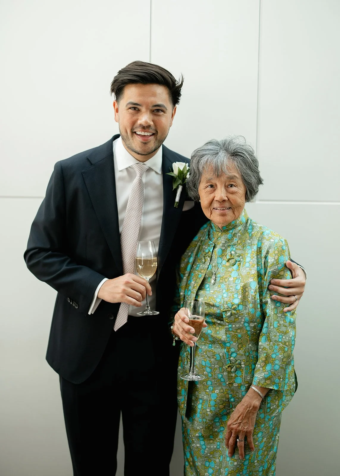 A young man in a suit with a boutonnière, holding a glass of champagne, standing next to an elderly woman in a colorful blouse, holding a glass of champagne, both smiling at the camera.
