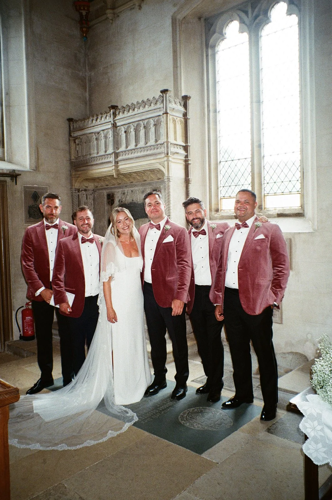 A bride in a white wedding dress with a long train, surrounded by six groomsmen in matching burgundy tuxedo jackets, white shirts, and burgundy bow ties, inside a historic church with tall stained glass windows.