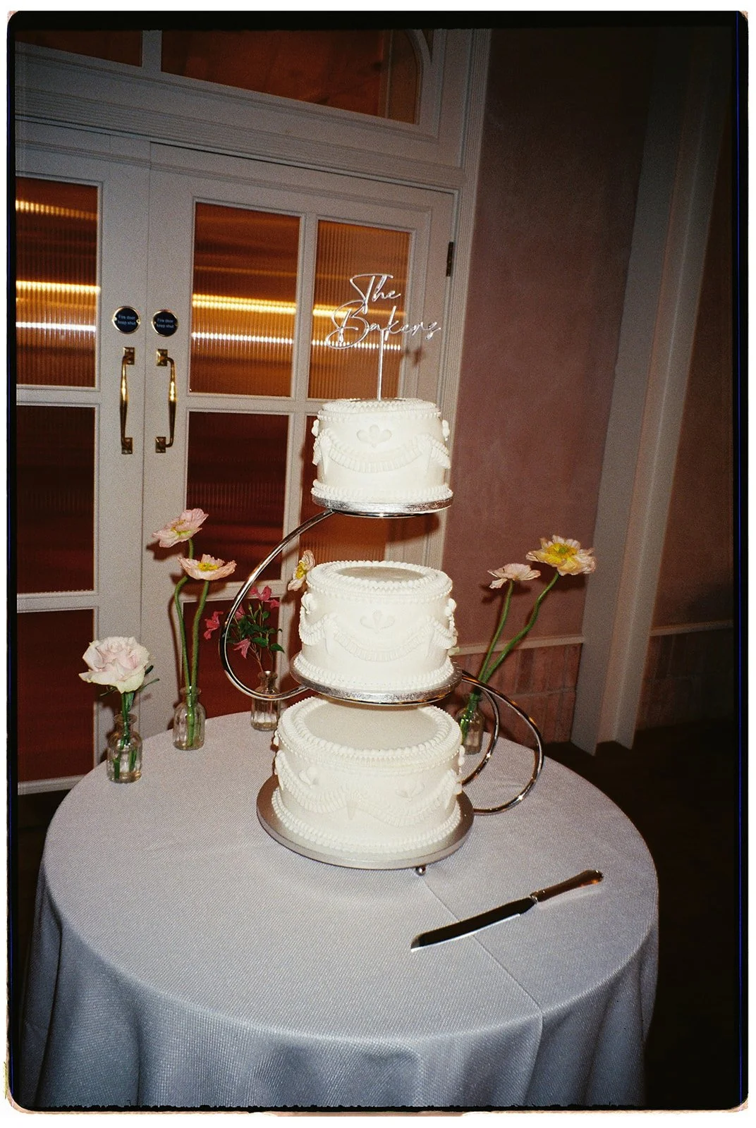 Three-tiered white cake on a table with pink flowers in vases nearby, a cake cutter, and a cake topper that says 'The Bakery' in cursive. The background features a set of double doors with frosted glass panels.