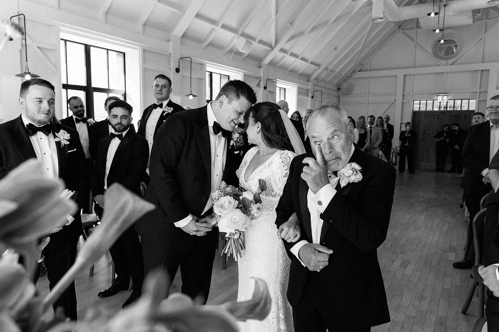 A black-and-white photo of a wedding ceremony with a bride and groom holding hands, leaning their foreheads together, surrounded by groomsmen and guests in a bright, airy room with large windows and wooden floors.