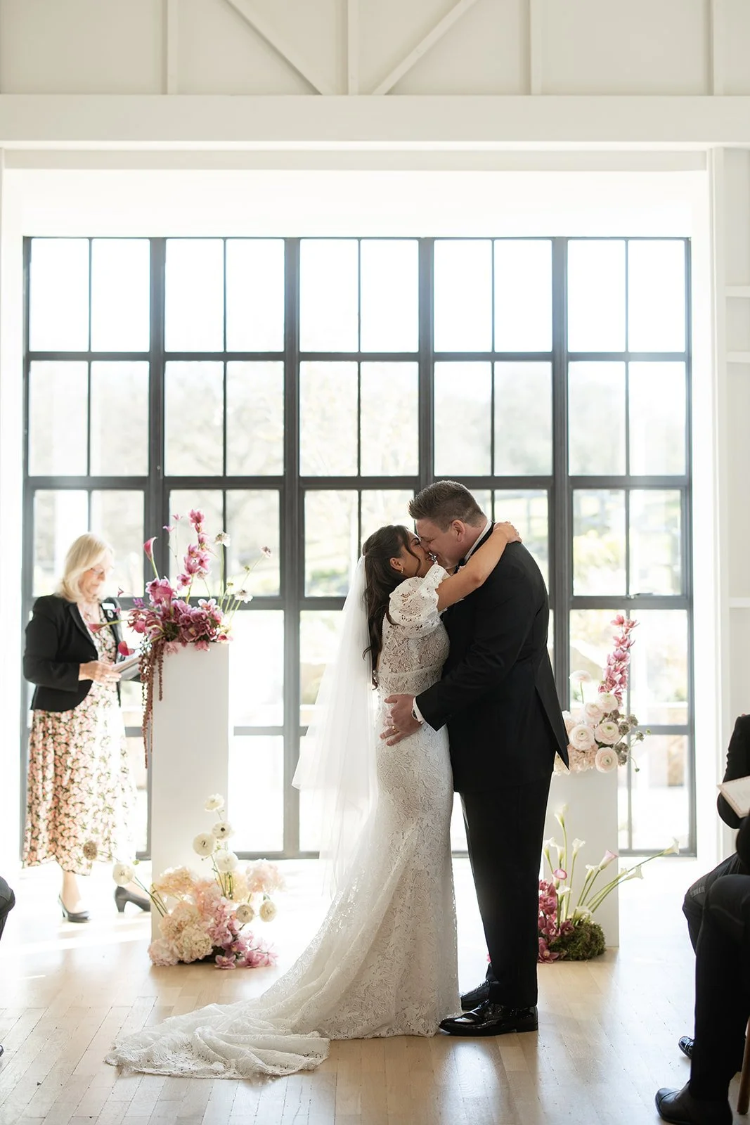 A bride and groom share a kiss during their wedding ceremony inside a bright room with large windows, decorated with pink and white flowers. An officiant or speaker is standing to the side, reading from a paper.
