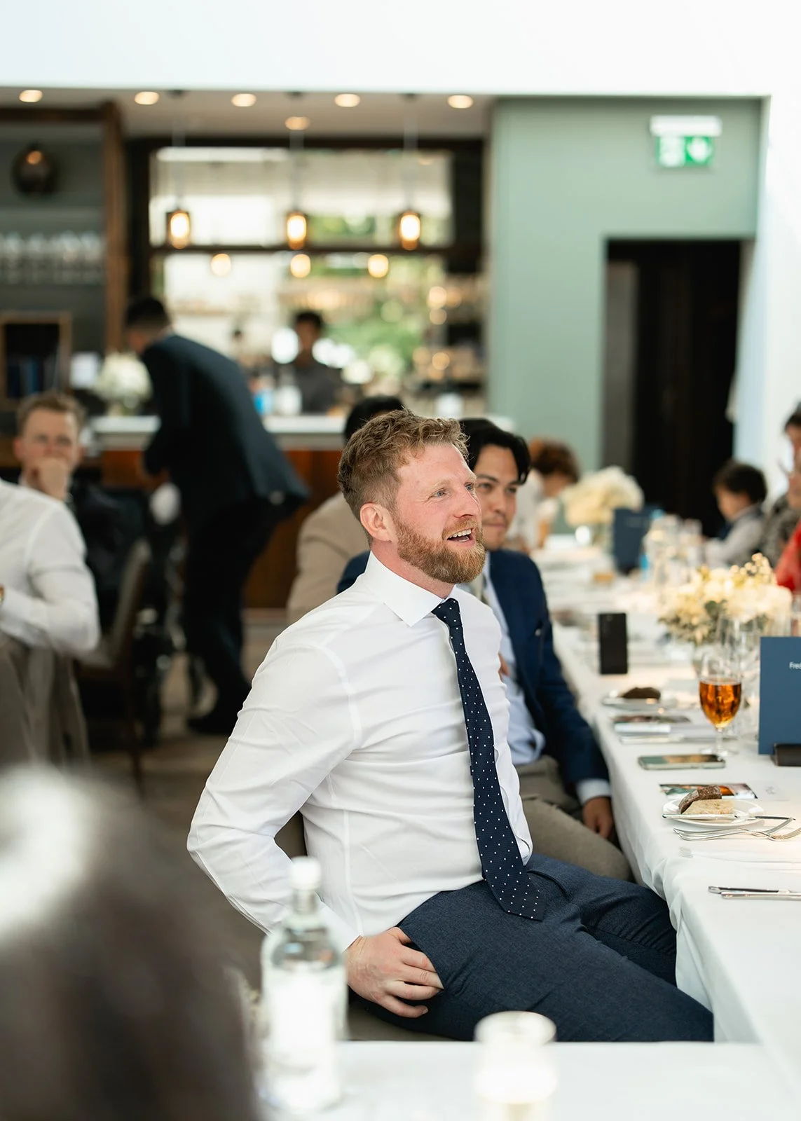 Man with a beard in a white shirt and navy polka dot tie sitting at a formal dinner event, smiling, with other suited guests and a server in the background.