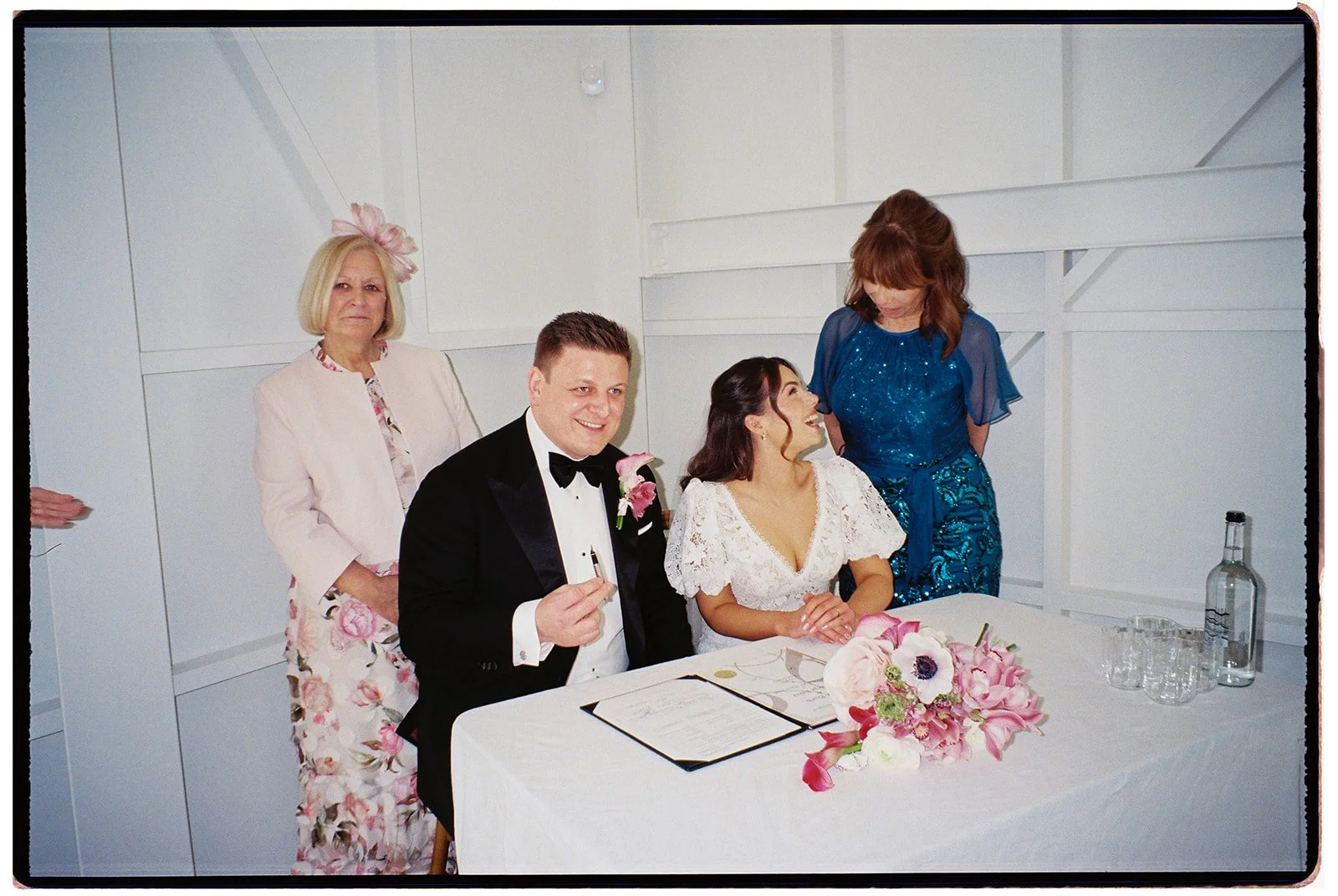 A wedding scene with a bride and groom seated at a table, surrounded by three women, one standing and two seated, in a white-walled room with floral decorations and a covered table with wedding documents and drinks.