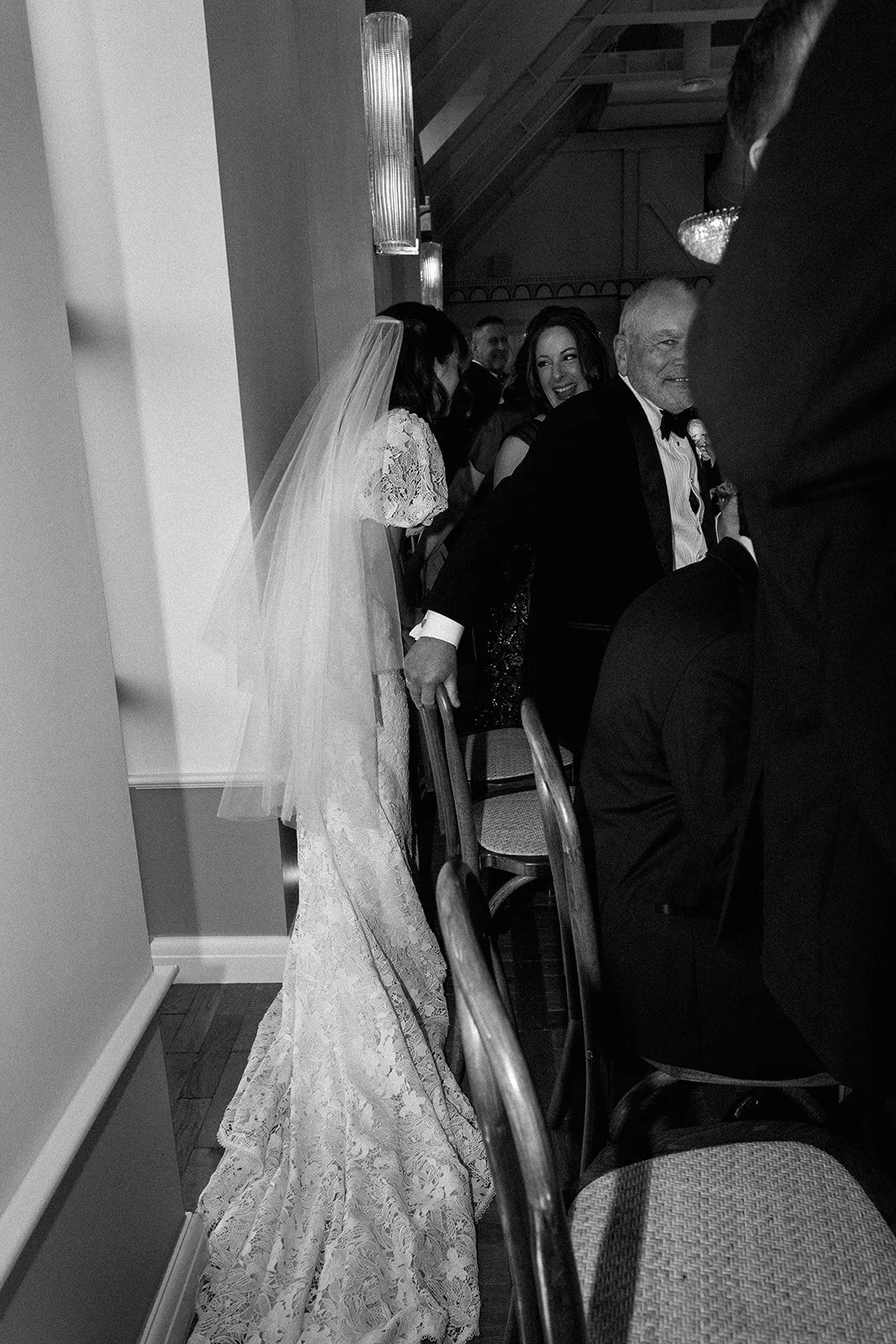 Wedding reception scene with a bride in a lace wedding dress and veil, smiling and leaning on a chair, surrounded by guests in formal attire.