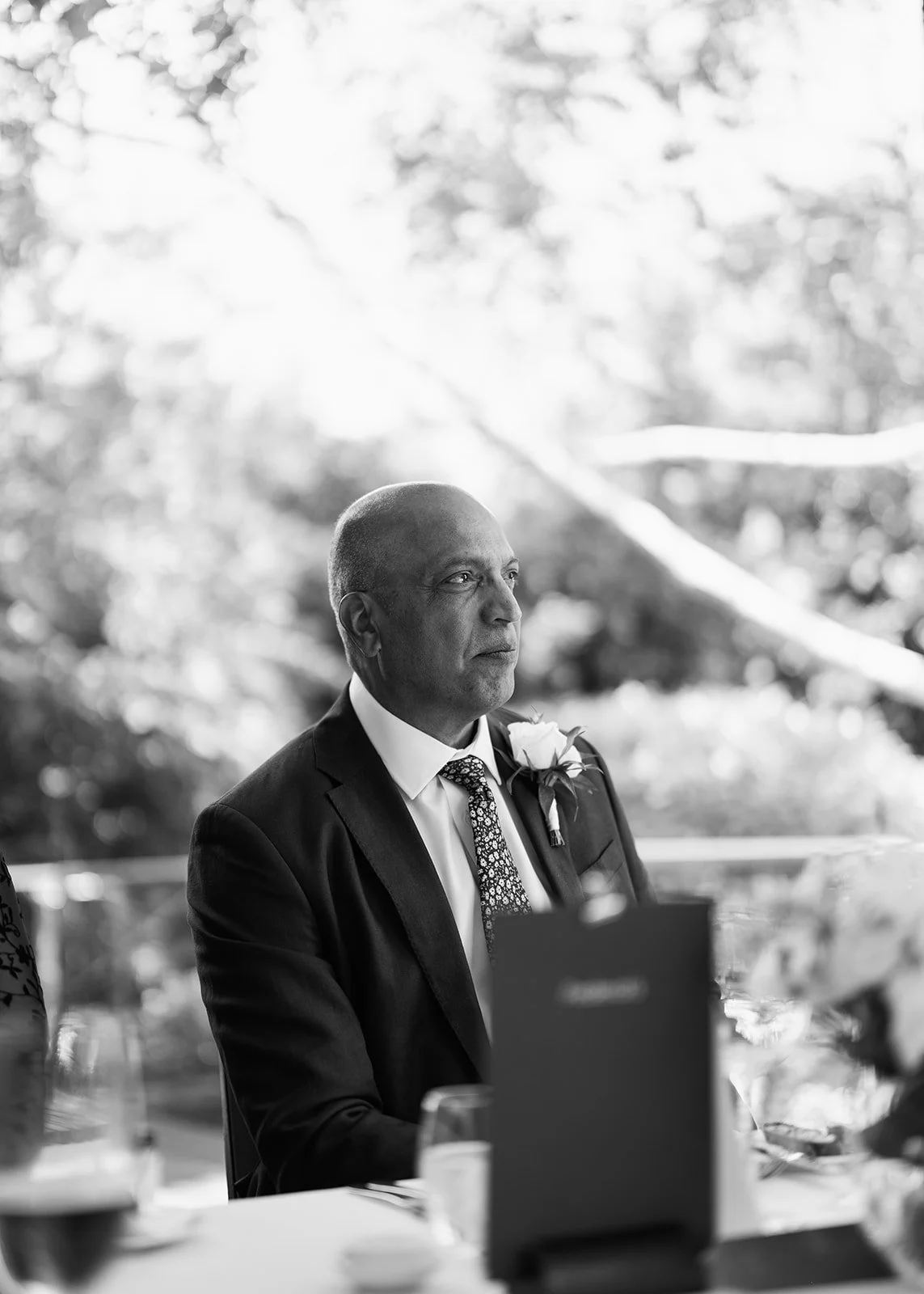 A black-and-white photo of a man in a suit with a boutonniere, sitting at a table during an outdoor event, looking contemplative.