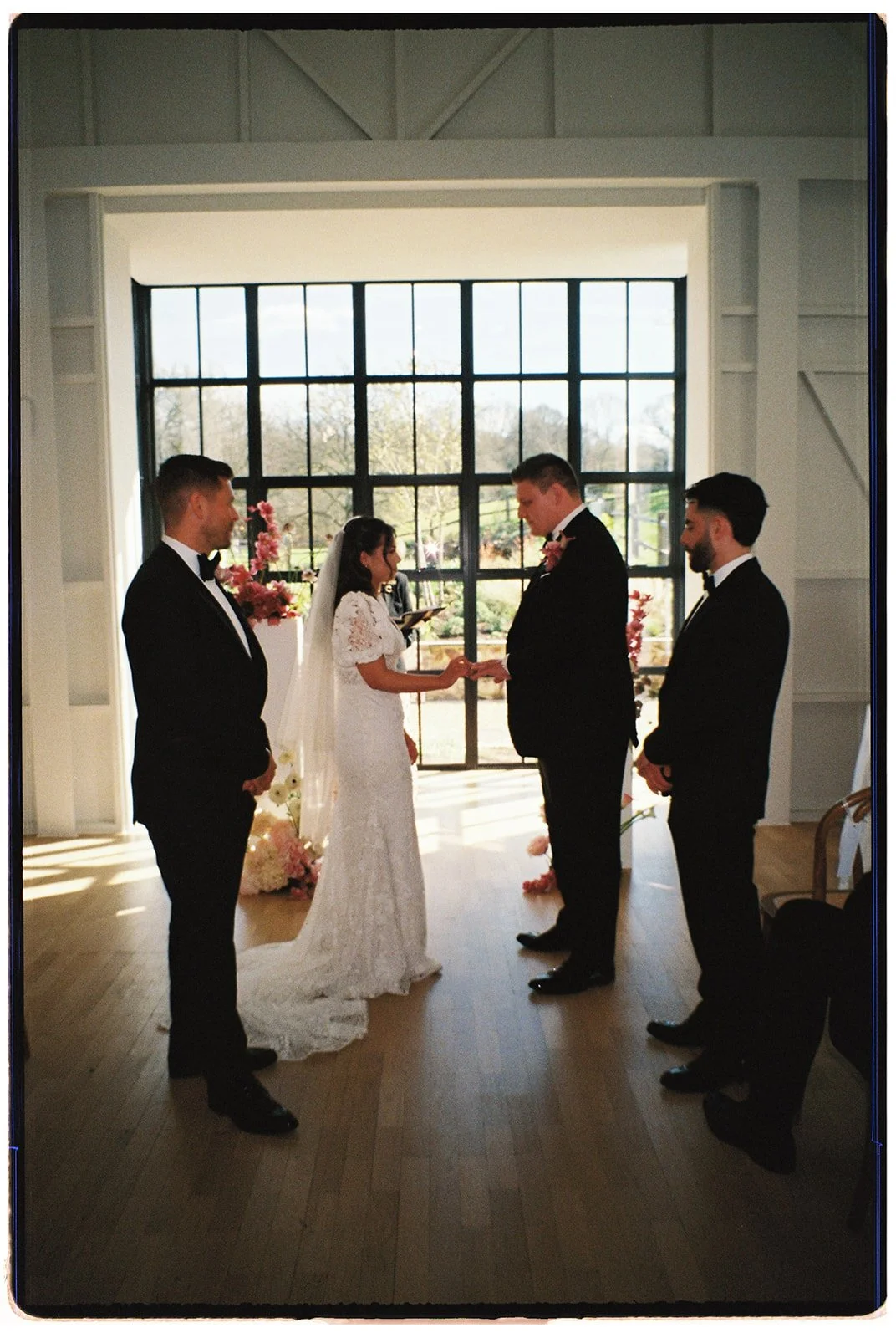 Bride and groom exchanging vows inside a bright room with large window, holding hands, flanked by two groomsmen in black tuxedos, with floral decorations nearby.