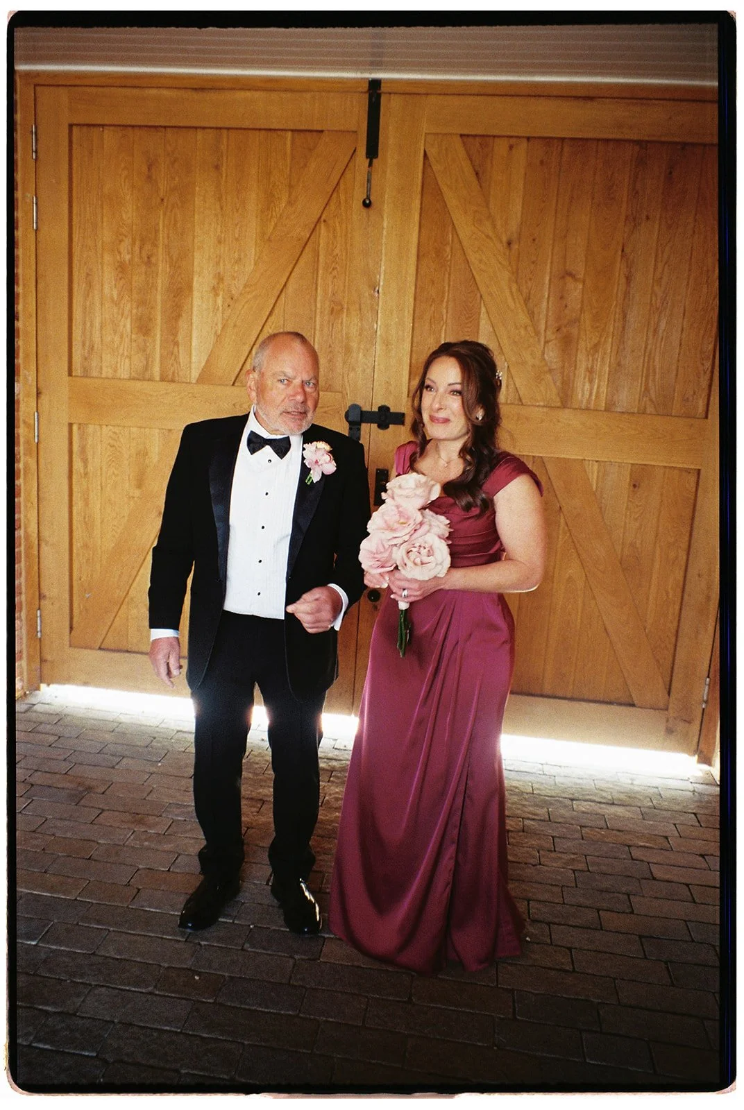 A man in a tuxedo and a woman in a maroon gown are standing in front of wooden barn doors. The woman is holding a bouquet of pink flowers and the man has a pink boutonniere. The man is making a gesture with his hand, and they are both smiling.