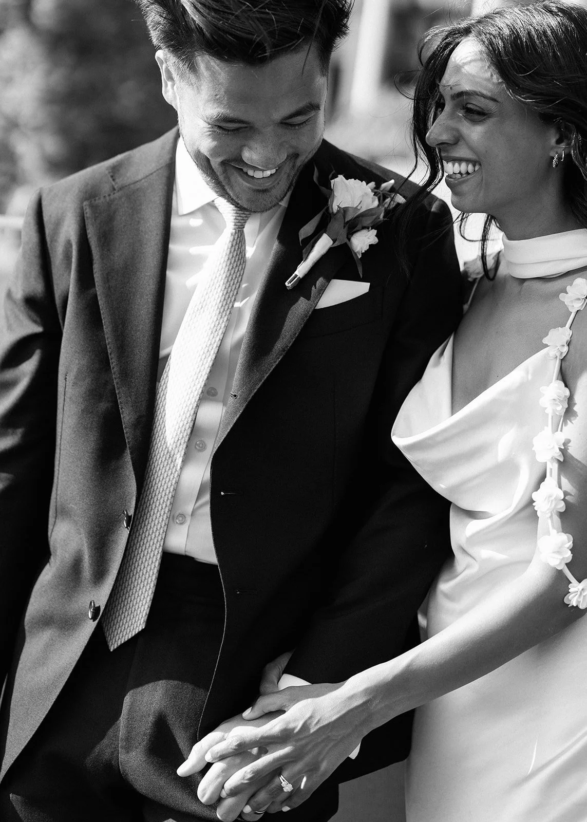 A black and white photo of a smiling couple holding hands, dressed in formal wedding attire, with the man in a suit and the woman in a wedding dress.