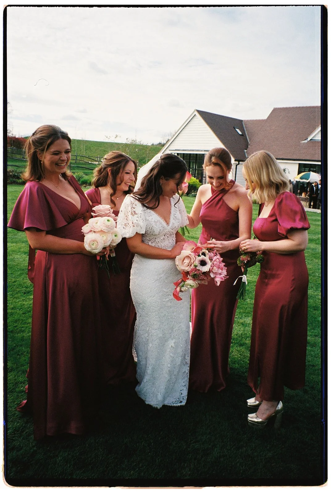 A bride in a white lace wedding dress surrounded by five bridesmaids in burgundy dresses, standing on a green lawn with a house in the background. The bride is holding a bouquet of pink and white flowers, and the bridesmaids are holding smaller bouqu