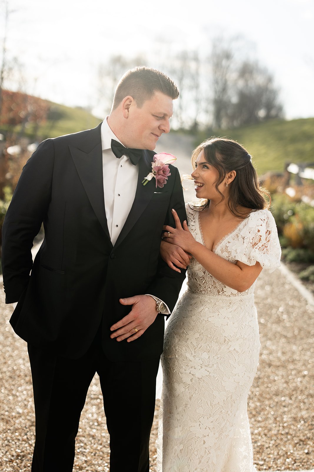 A bride and groom stand together outdoors, smiling at each other. The groom is dressed in a black tuxedo with a purple boutonniere, and the bride is wearing a lace wedding dress, holding his arm. Sunlight and a blurred natural background are visible.