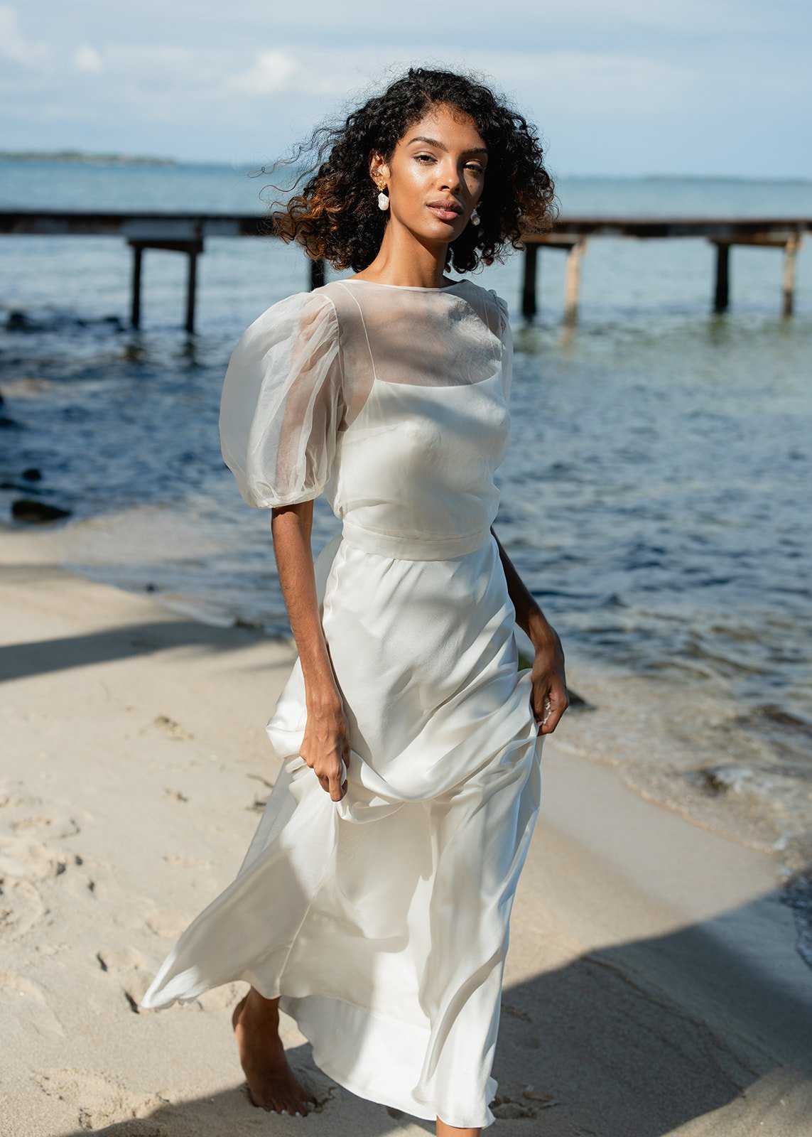 A woman in a white dress walking barefoot on a sandy beach near the ocean, with a wooden pier in the background.