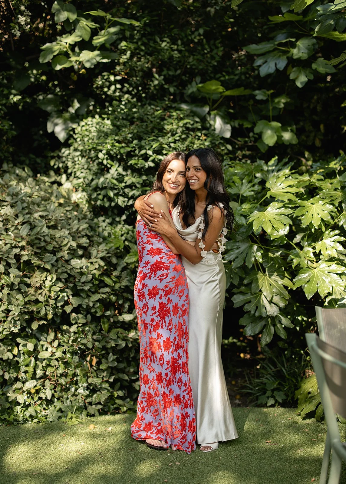 Two women dressed in elegant dresses standing closely and hugging each other in a lush green garden.