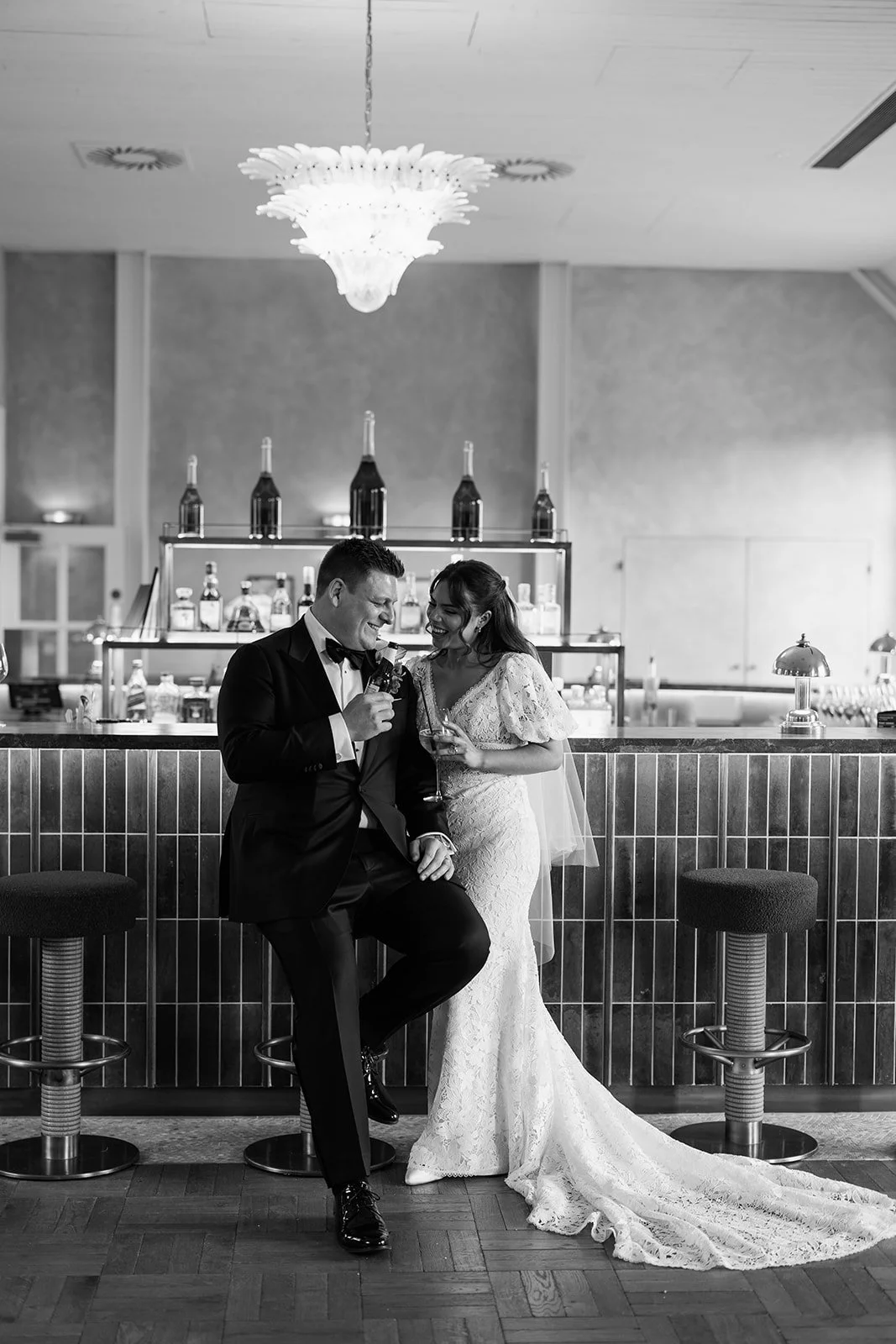 A newlywed couple in wedding attire sharing a moment at a bar at Limeklin, with the groom in a tuxedo and the bride in a lace wedding dress, holding glasses, in a modern indoor setting.