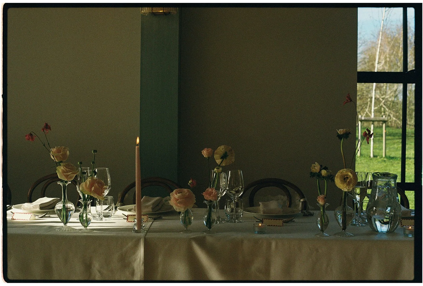A dining table set for a formal meal with vases of flowers, wine glasses, and a tall candle, situated near a window showing an outdoor scene with trees and grass.