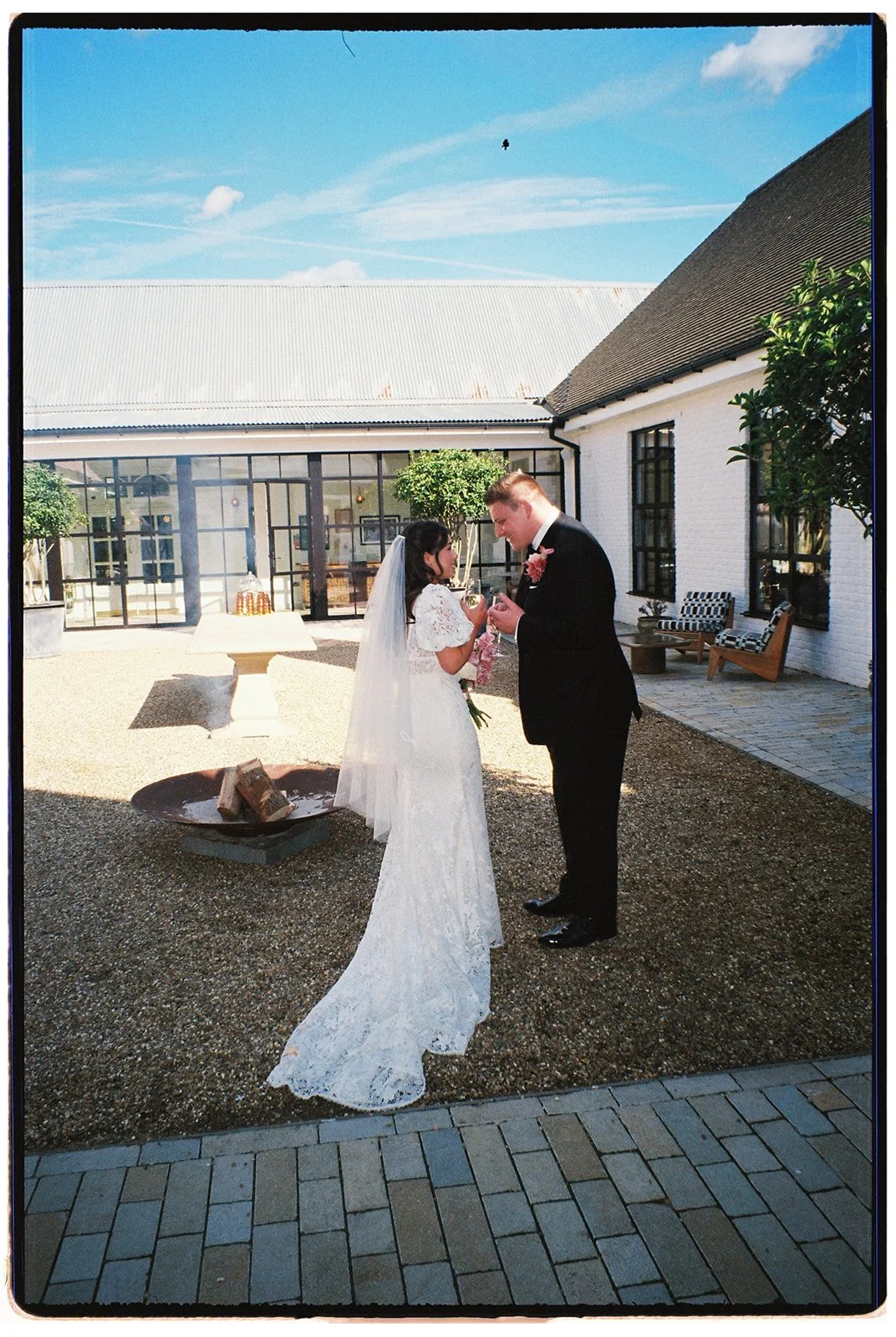 A bride in a white lace wedding gown and veil, holding a pink bouquet, and a groom in a black tuxedo, sharing a moment and a cigar outside a wedding venue with modern windows and outdoor seating.