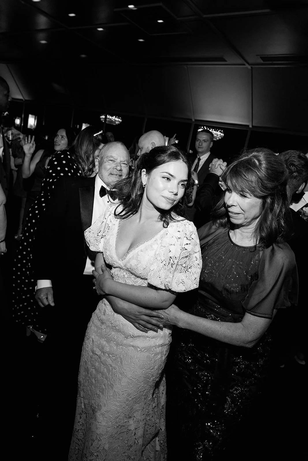 A black-and-white photo of a woman in a lace dress dancing with another woman at a formal event, surrounded by other guests.
