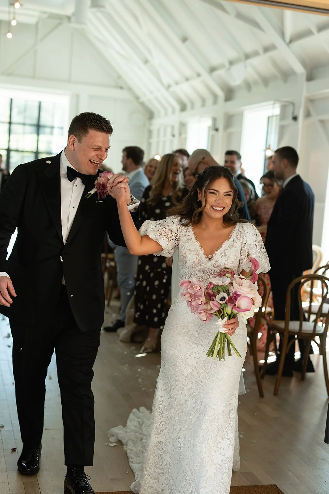 Bride and groom smiling and holding hands at their wedding reception, surrounded by guests in a bright, white, rustic venue.