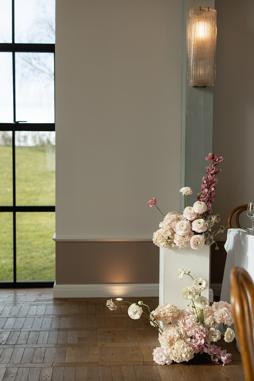 Interior view of a room with a large window, a wall-mounted light fixture, and a floral arrangement in a white vase containing pink and white flowers on the floor near a table.