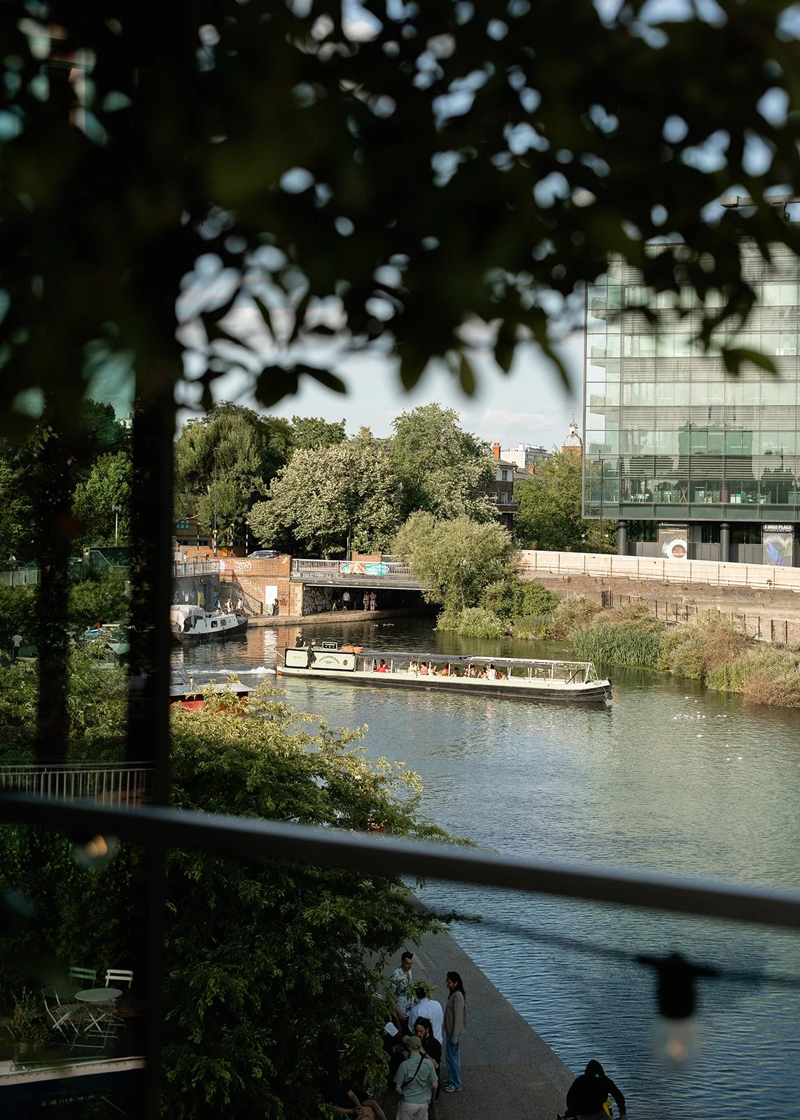 A city canal scene with a boat carrying several passengers, surrounded by green trees and modern buildings. People are gathered along the water's edge, and a bridge spans the canal in the background.