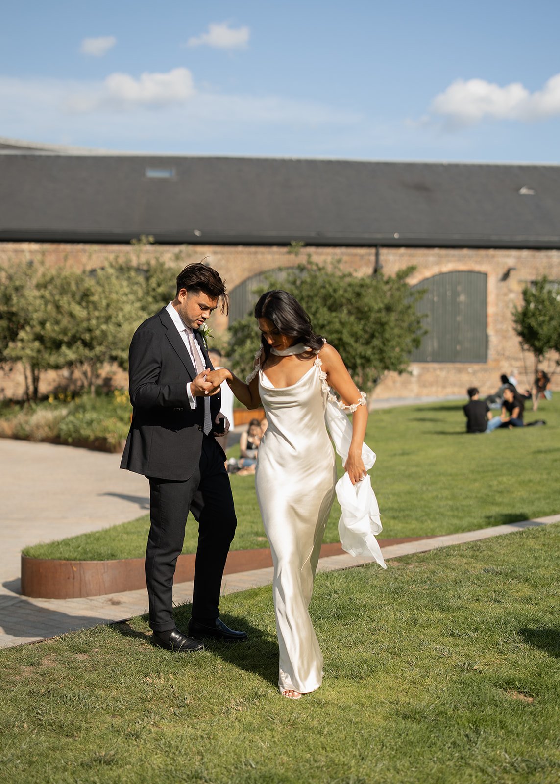 A bride and groom walking on grass in an outdoor location. The bride is wearing a white dress and the groom is dressed in a black suit. The groom is holding the bride's hand while they walk.