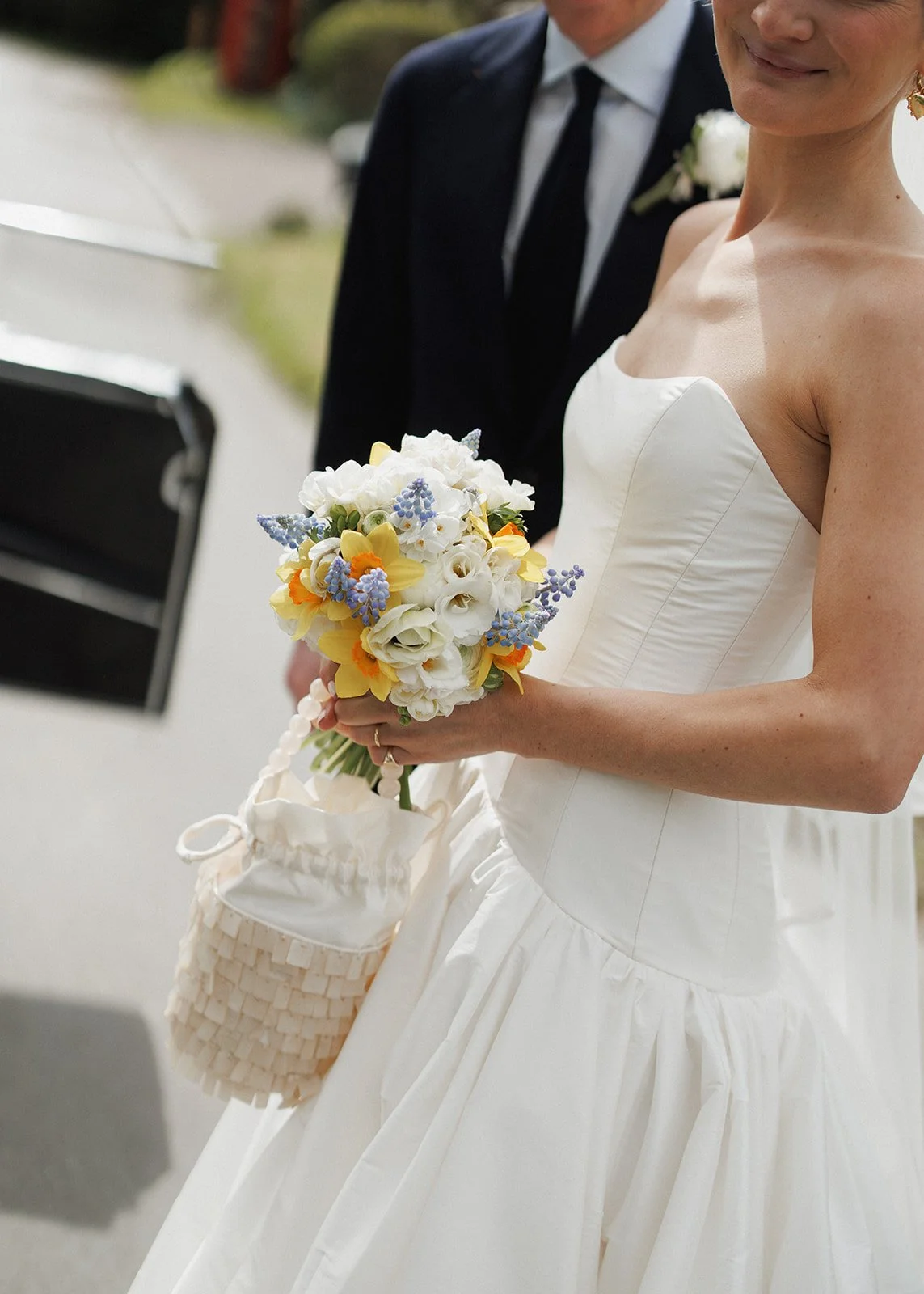 A bride in a white strapless wedding dress holding a bouquet of white, yellow, and purple flowers, with a small woven basket hanging from her arm, standing outdoors with a groom in a black suit and tie in the background.