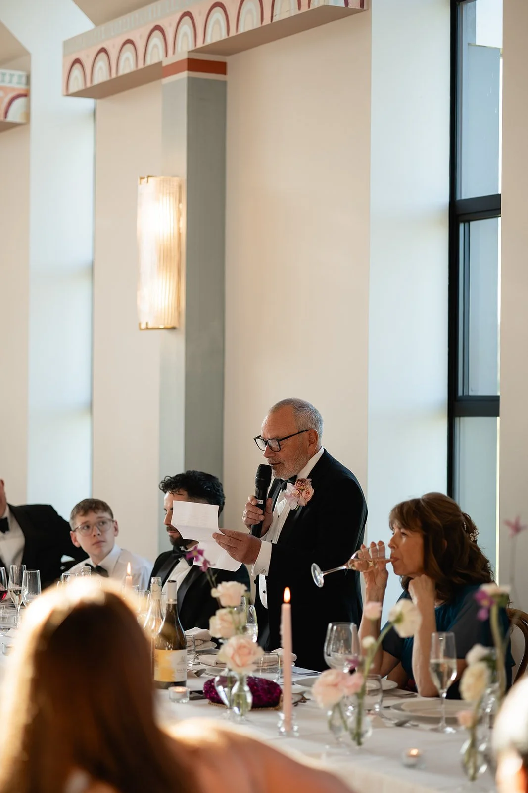 An elderly man in a tuxedo giving a speech at a wedding reception, holding a paper and a microphone, surrounded by guests seated at a decorated table with flowers and candles.