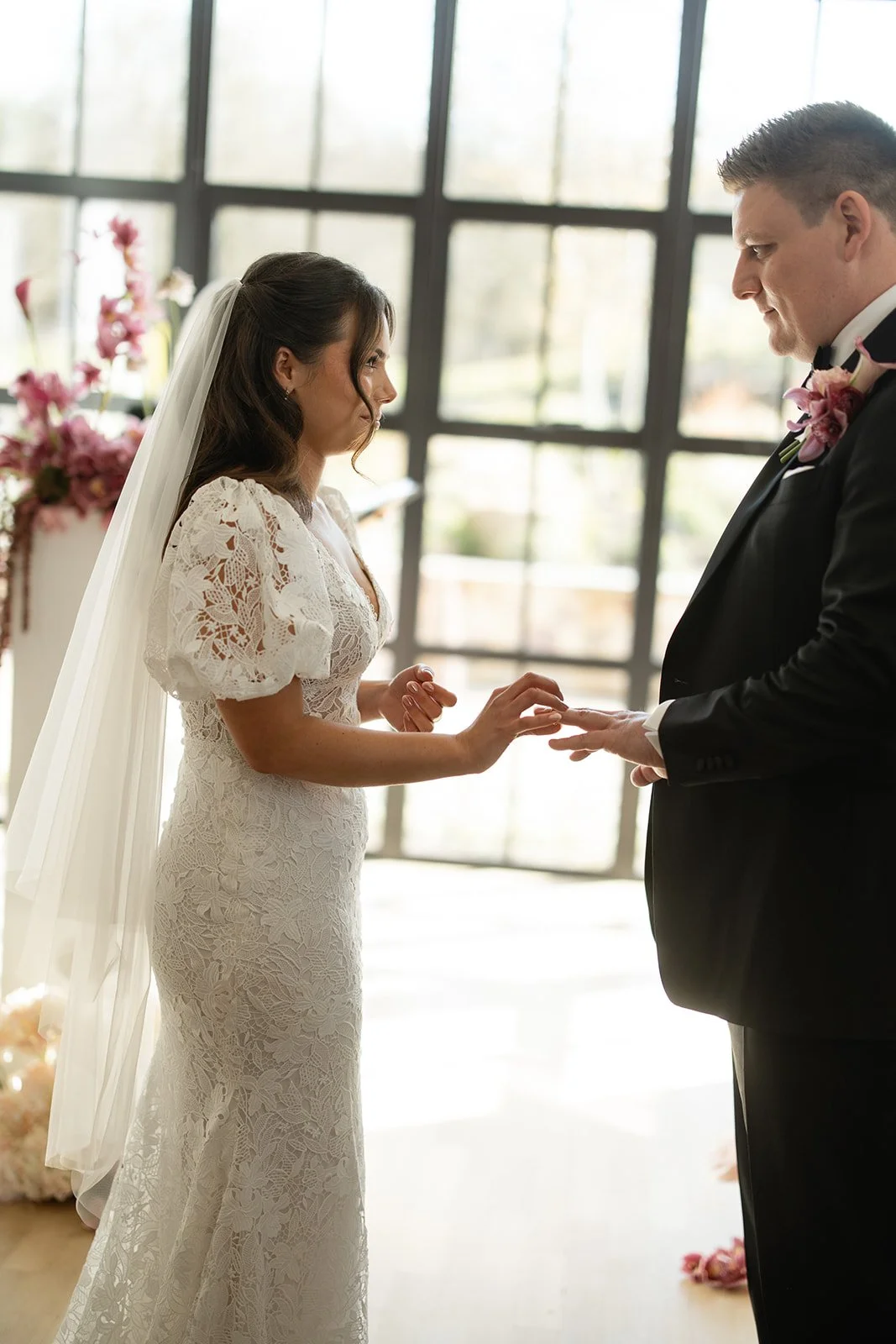 A bride and groom exchanging wedding rings indoors with large windows and pink floral decorations in the background.