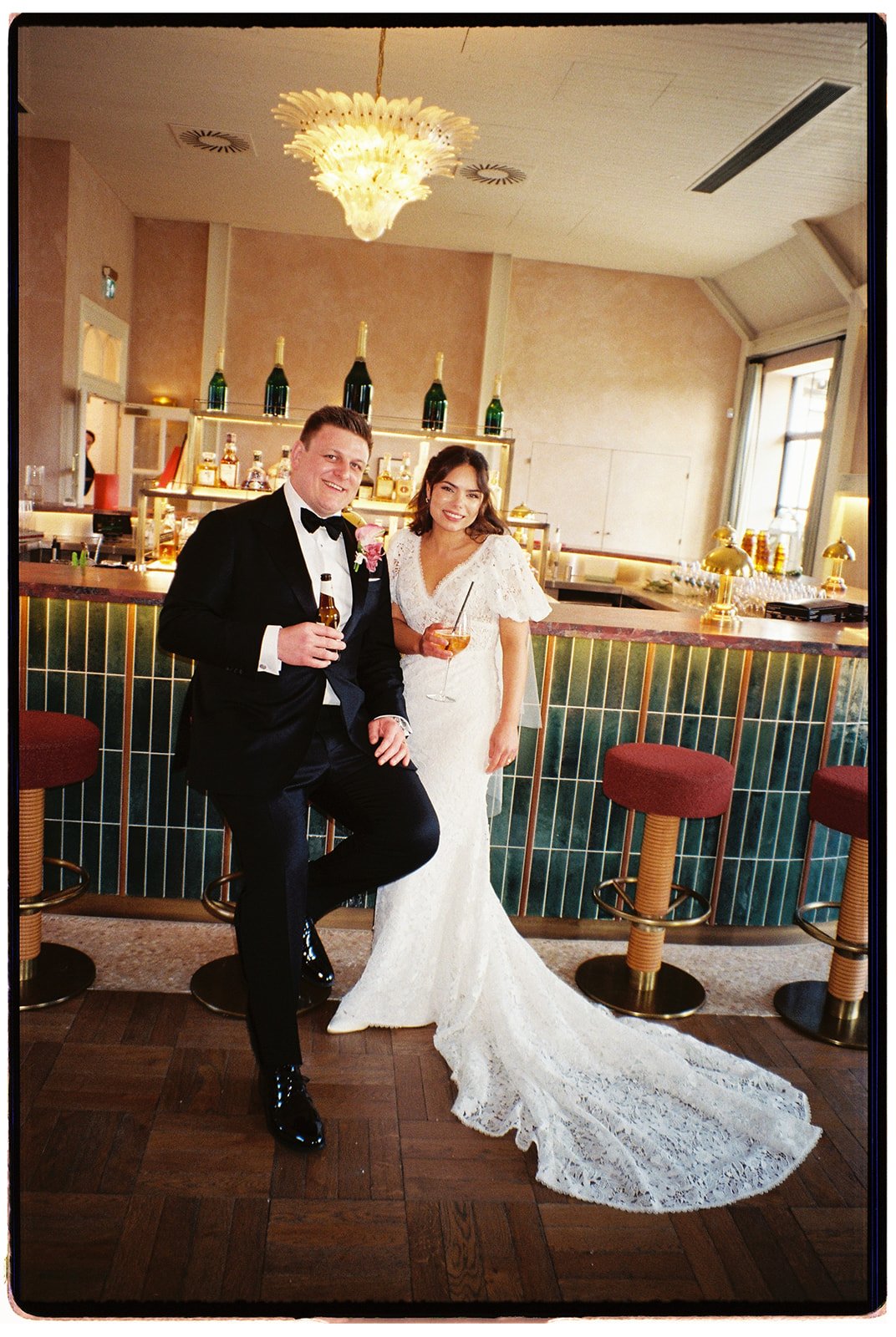 A bride and groom at a wedding reception, smiling and holding drinks, standing in front of a bar with green tiles and bottles on shelves, decorated with a large ornate chandelier and gold accents.