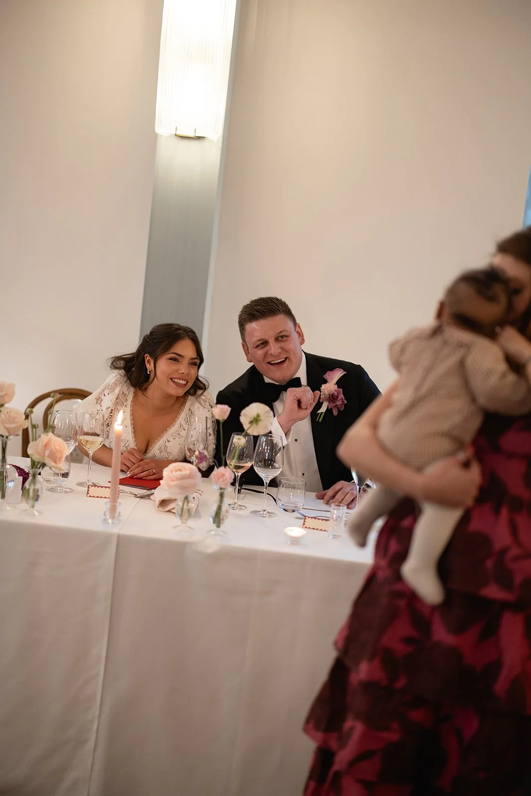A wedding reception with a bride, groom, and a woman holding a small child at a decorated table with flowers and candles.
