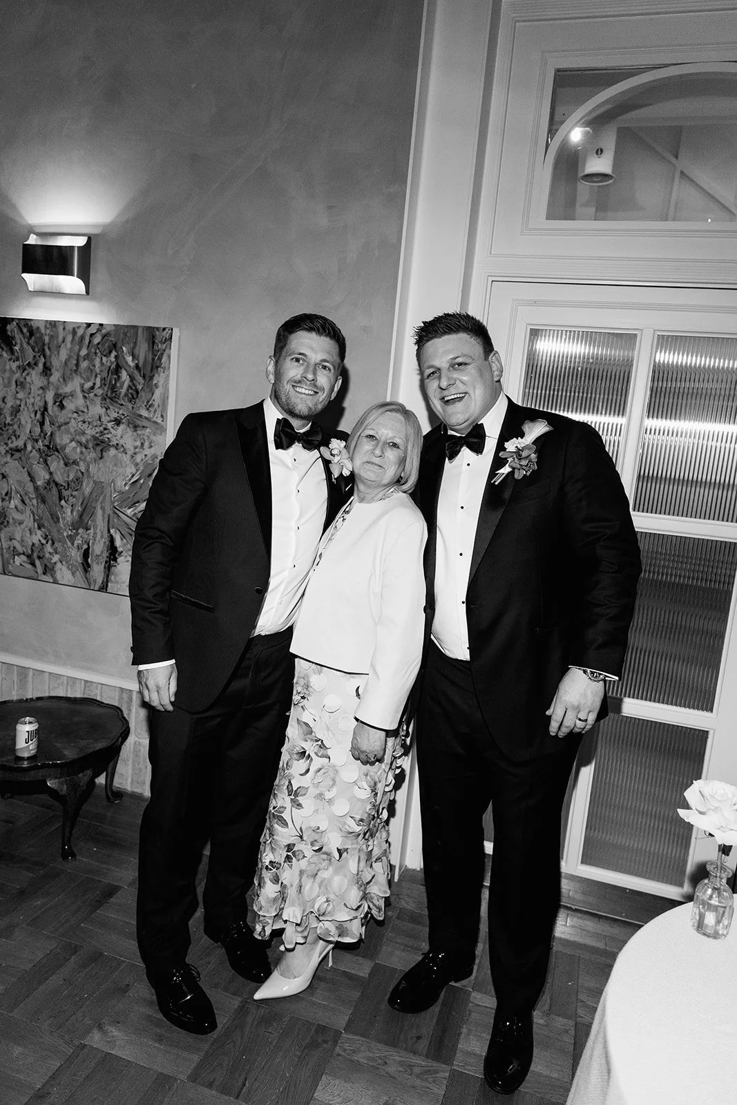 Three people in formal attire smiling at a social event, two men in tuxedos and a woman in a floral skirt and white top, standing close together in a well-lit room.