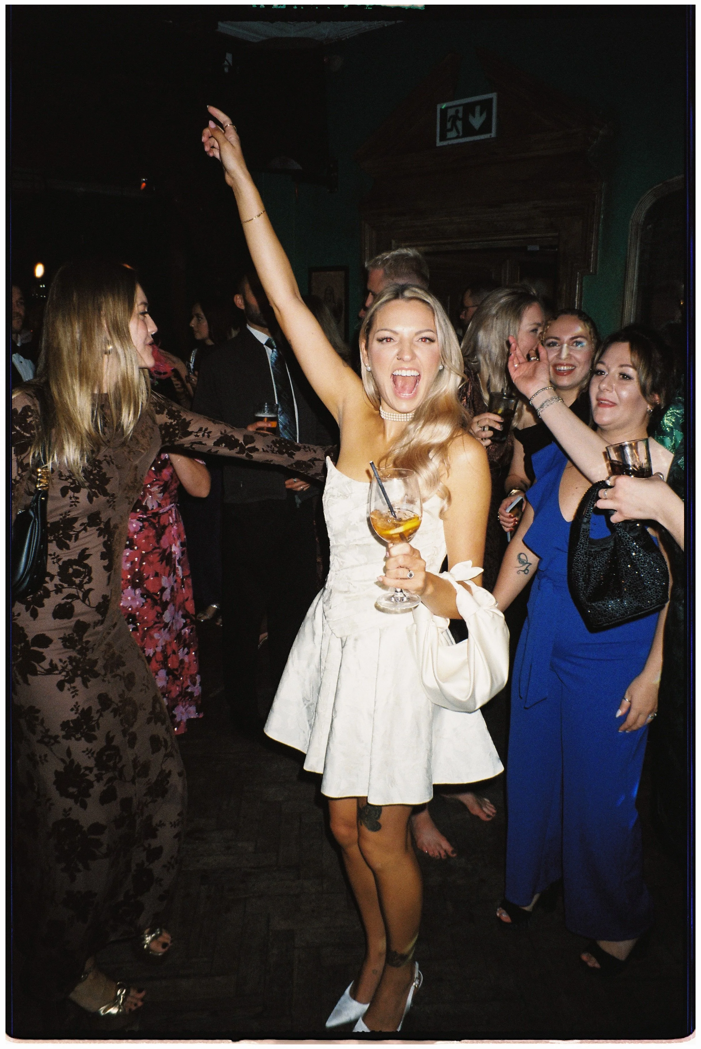 A group of women enjoying a party or celebration, with one woman in a white dress holding a drink and cheering enthusiastically, surrounded by friends.