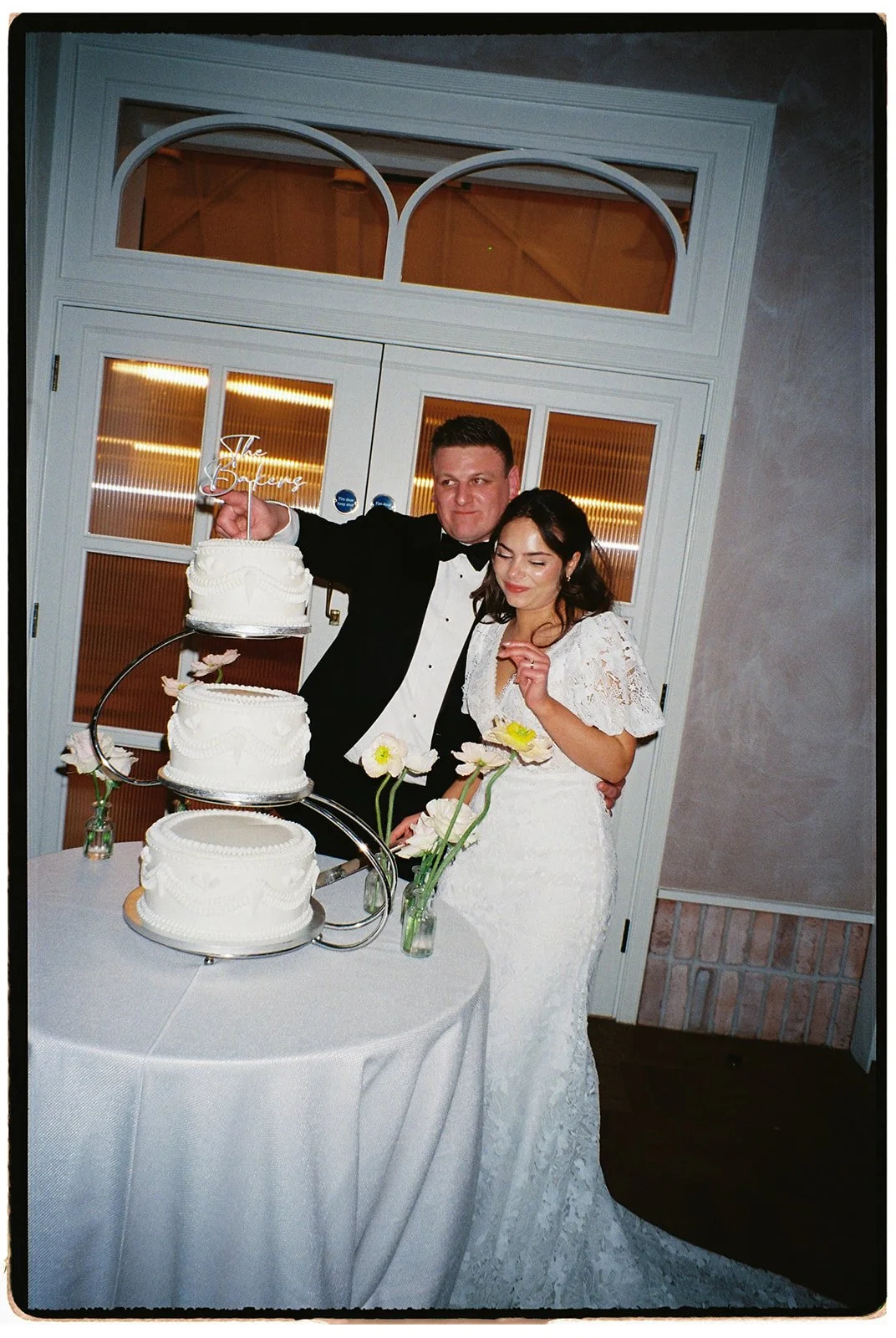 A bride and groom cutting a three-tiered wedding cake together at their wedding reception, with flowers on the table and a sign on top of the cake that says 'The Best is Yet to Come'.