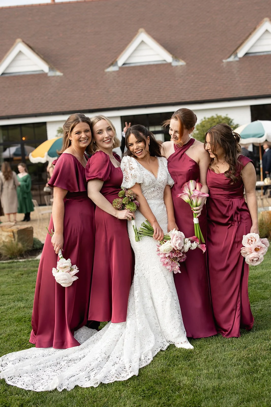 Group of women at a wedding, with the bride in a white lace dress holding a bouquet of pink and white flowers, standing outdoors with a house and guests in the background.