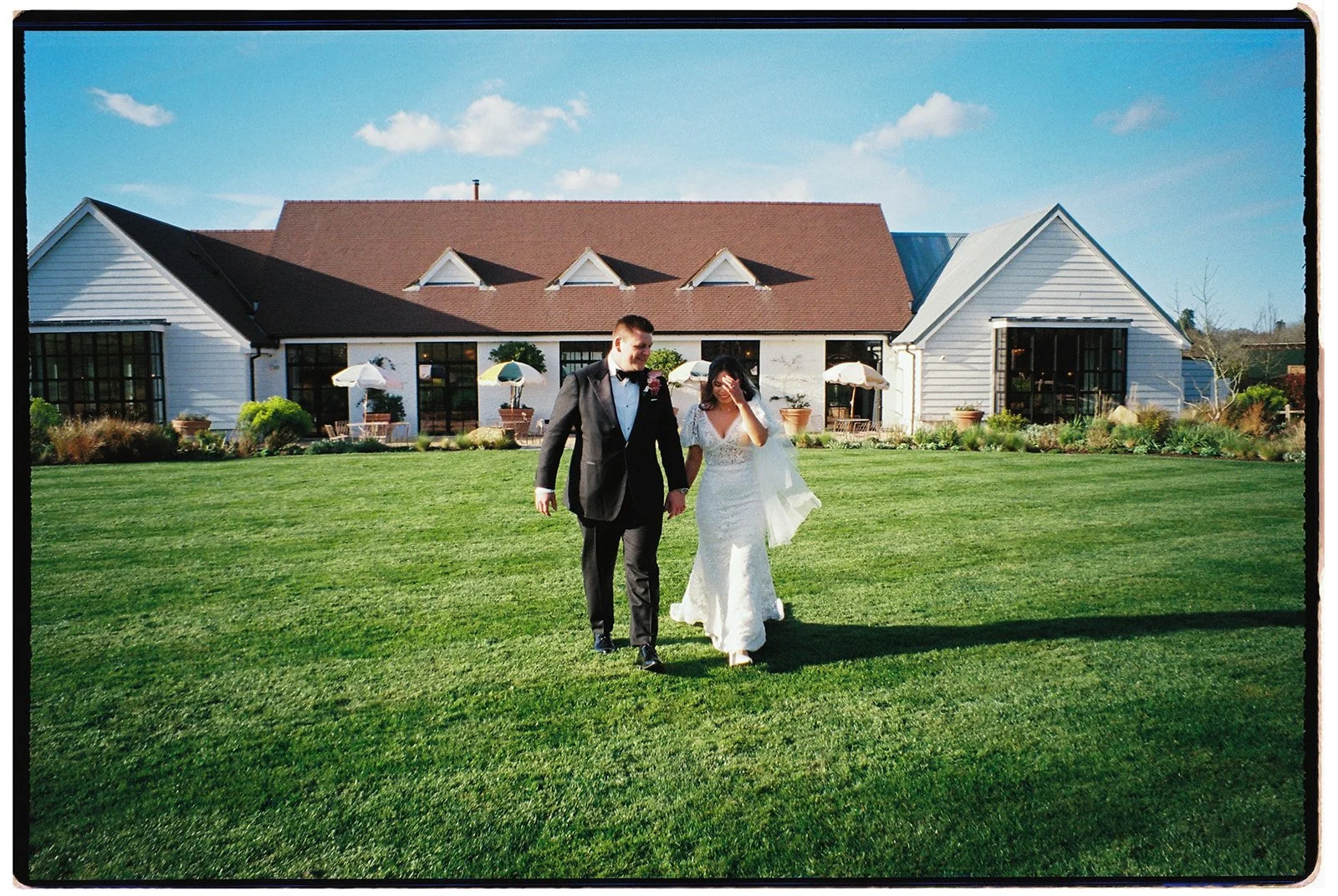 A bride and groom walking on a grassy lawn outside a white wedding venue, with umbrellas and potted plants in the background on a sunny day.