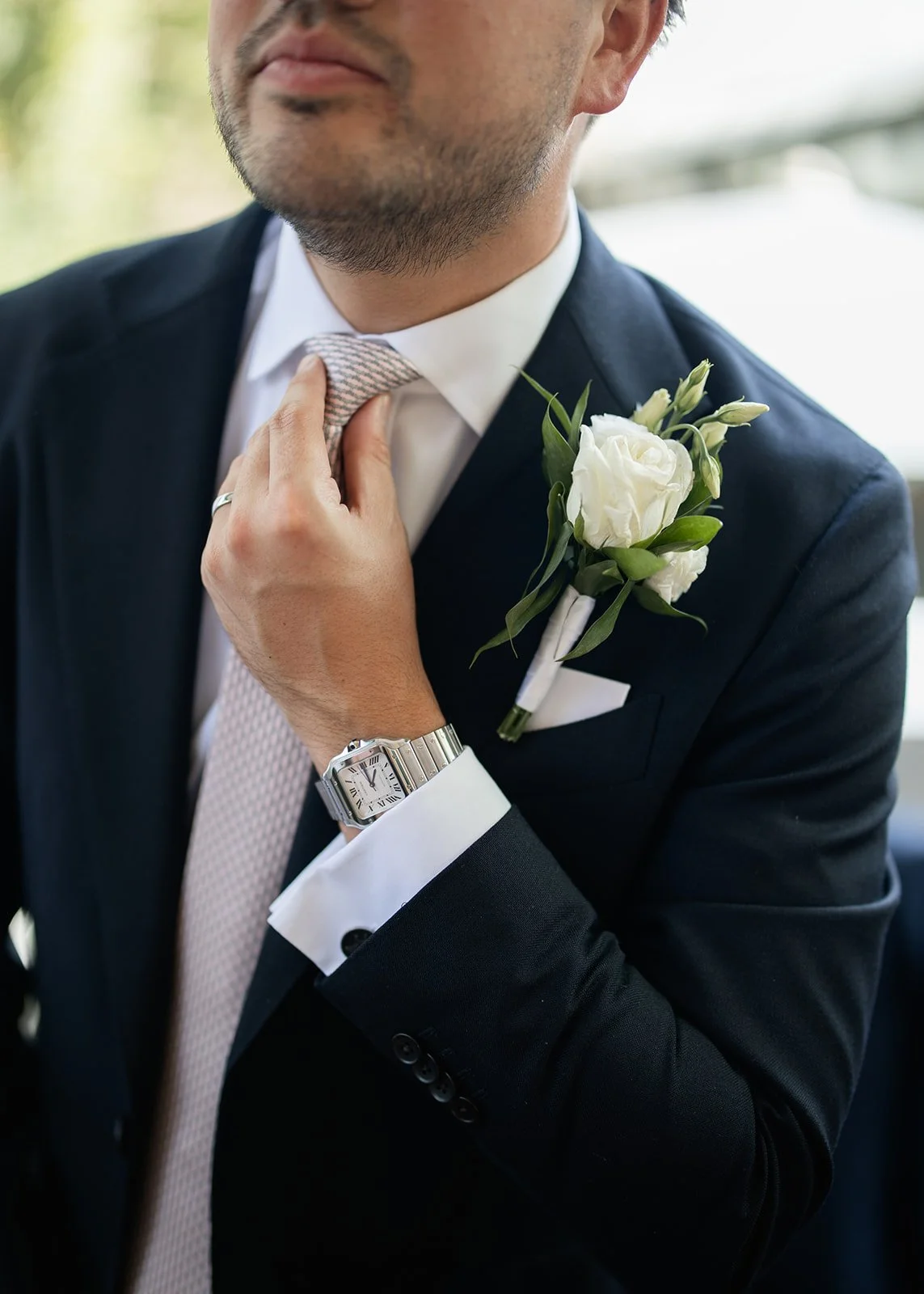 A man dressed in a black suit with a white shirt, adjusting his tie. He is wearing a wristwatch and a wedding ring. A white flower boutonniere is pinned to his lapel.