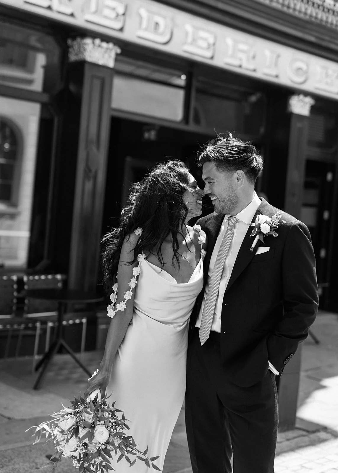 A black-and-white photo of a wedding couple sharing a moment. The bride is holding a bouquet and wearing a dress with floral details on the shoulders, while the groom is in a suit with a boutonniere. They are smiling and touching foreheads in front of a storefront.