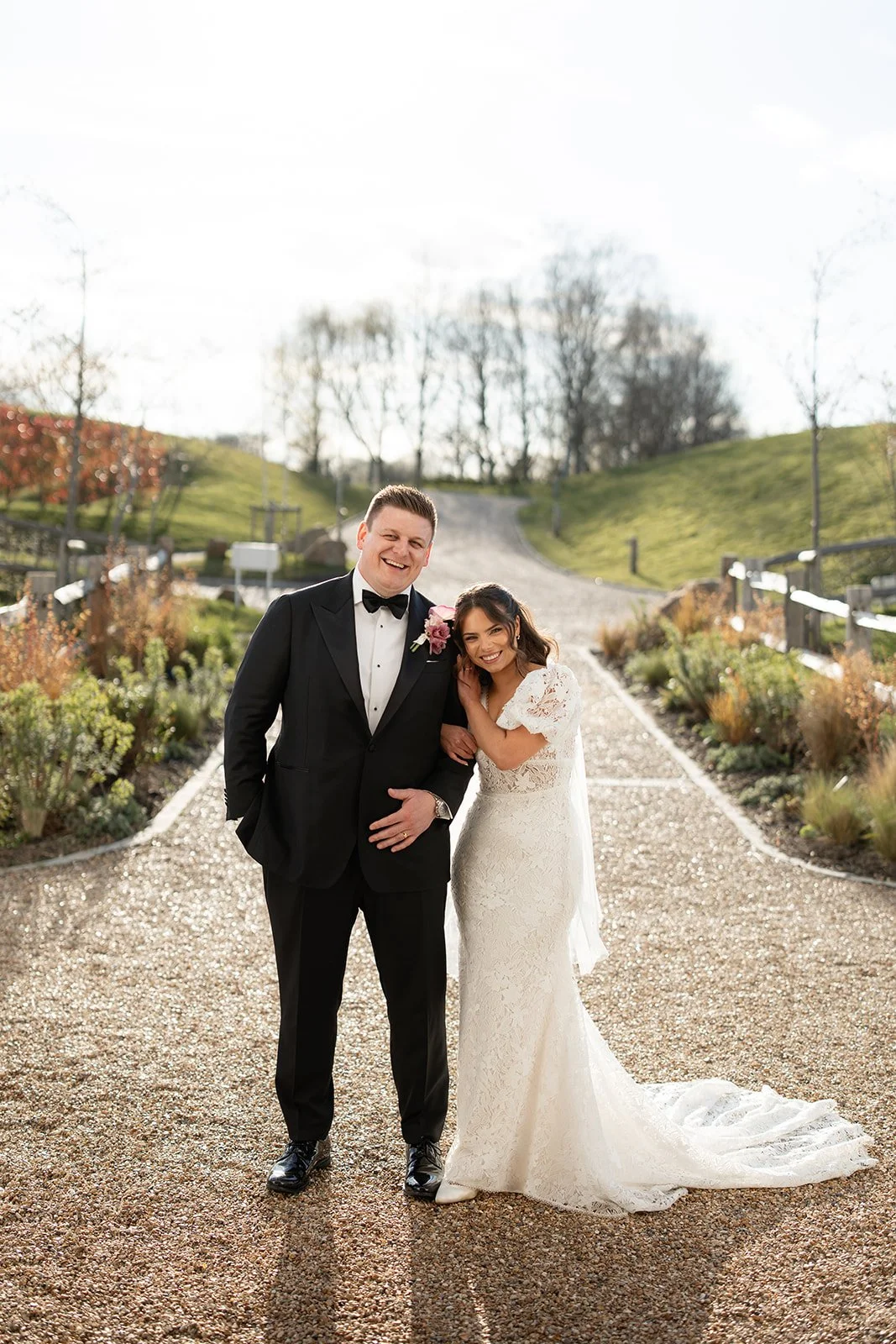 A smiling couple dressed in wedding attire standing on a pathway outdoor side LIMEKILN, with autumn trees and a hill in the background.