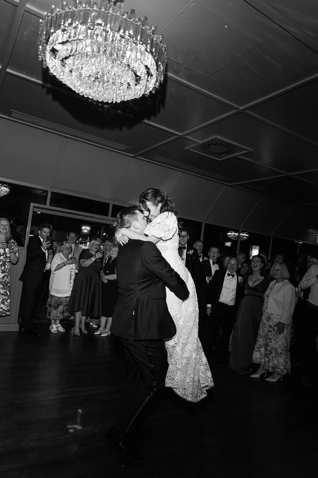 A bride and groom dancing at their wedding reception, surrounded by guests in formal attire, with a chandelier hanging from the ceiling.