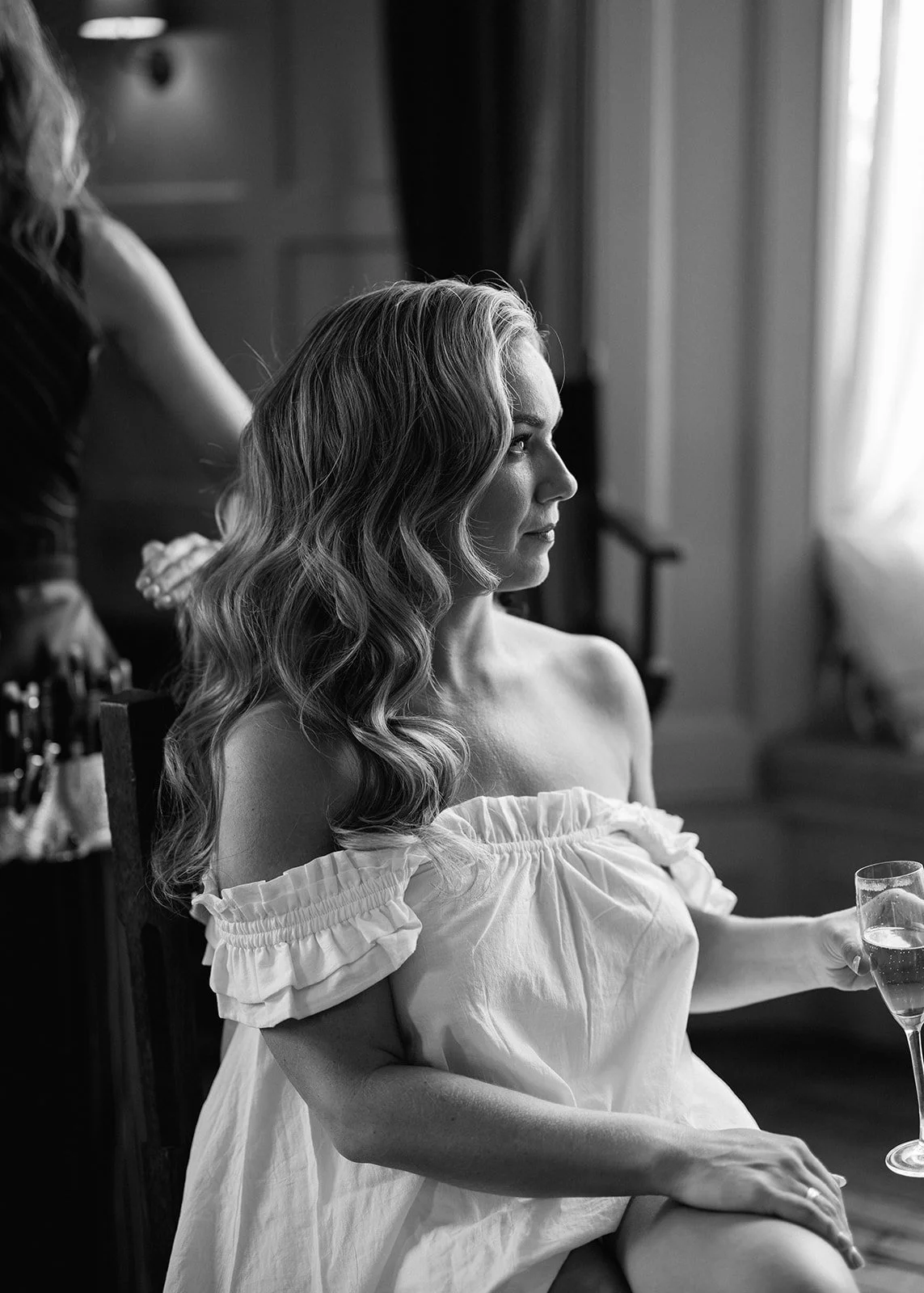 A woman with wavy hair sitting on a chair, holding a glass of champagne, wearing an off-the-shoulder dress, with a woman standing behind her in a room with natural light and curtains.