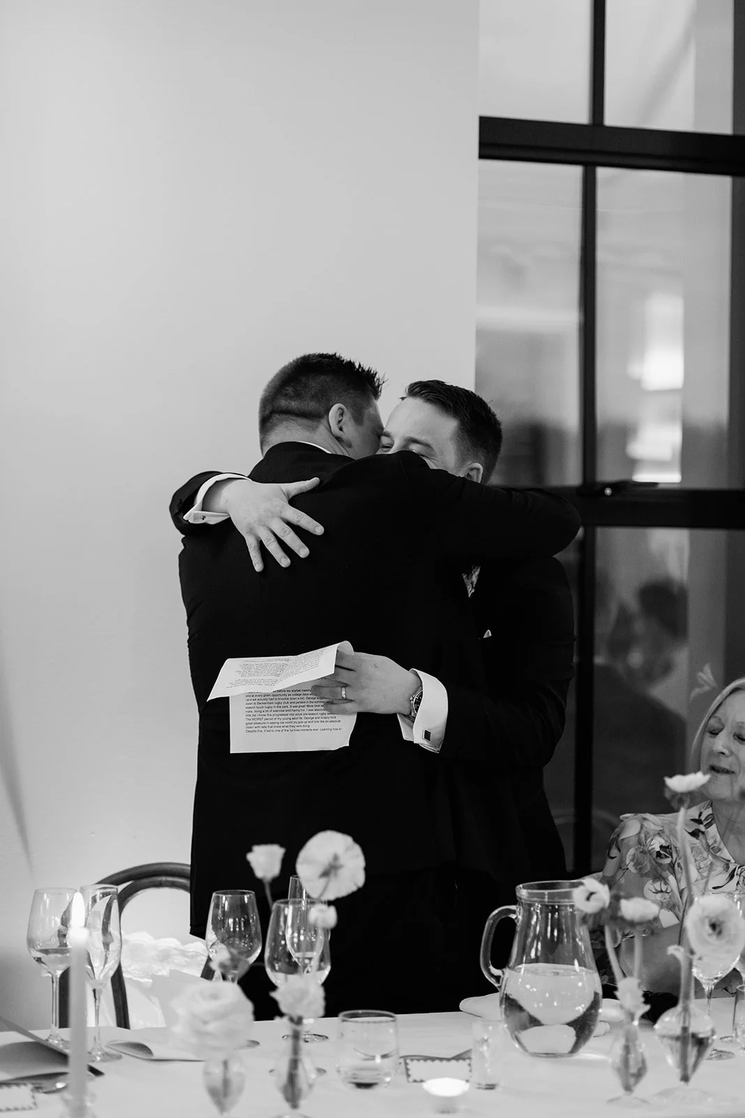 Two men in suits sharing a hug at a formal event, with a woman sitting at a table with flowers and glasses in the foreground.