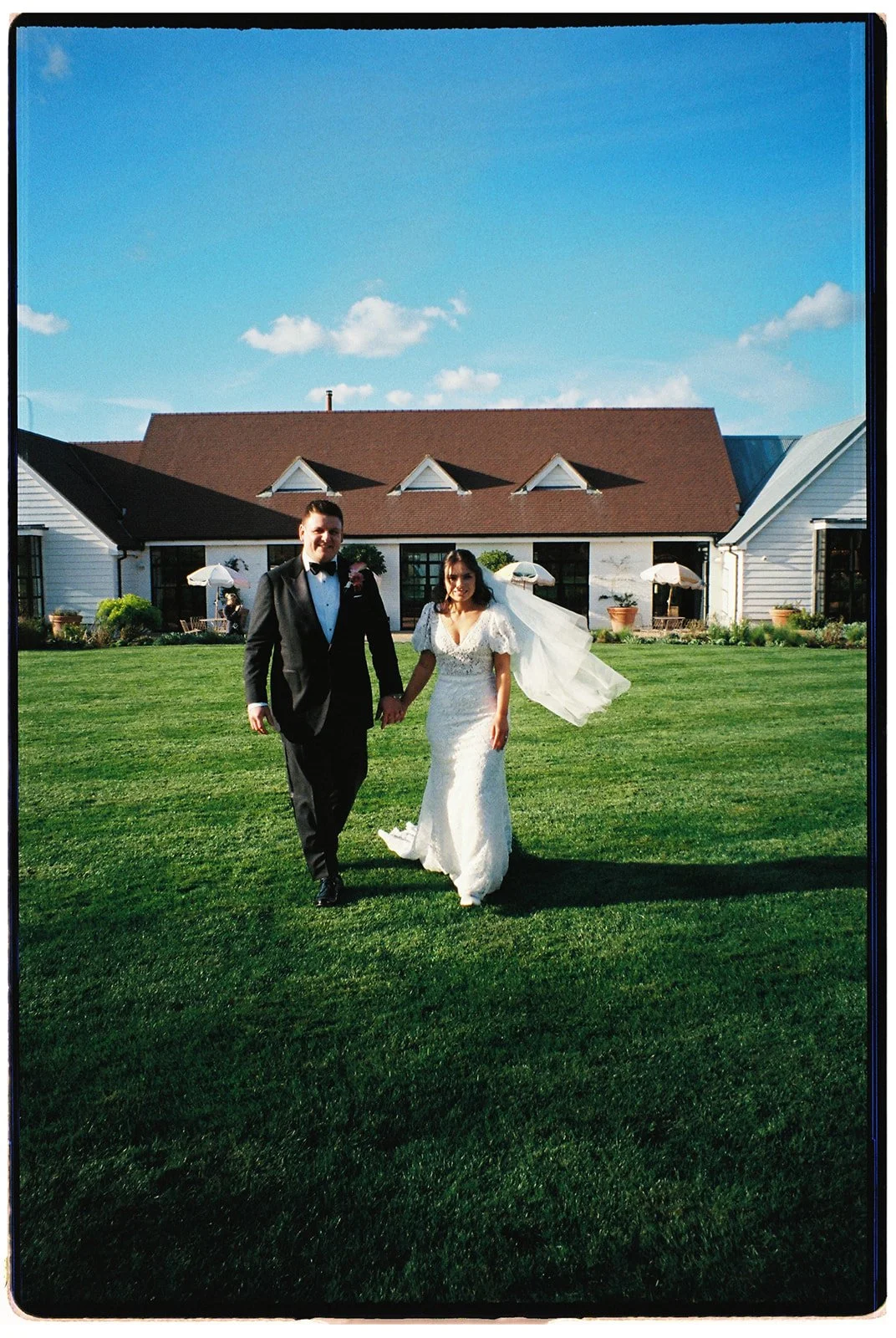 A bride and groom holding hands and walking on a well-kept lawn outside a white house with a brown roof on a sunny day. The bride is wearing a white wedding dress and veil, and the groom is in a black tuxedo.