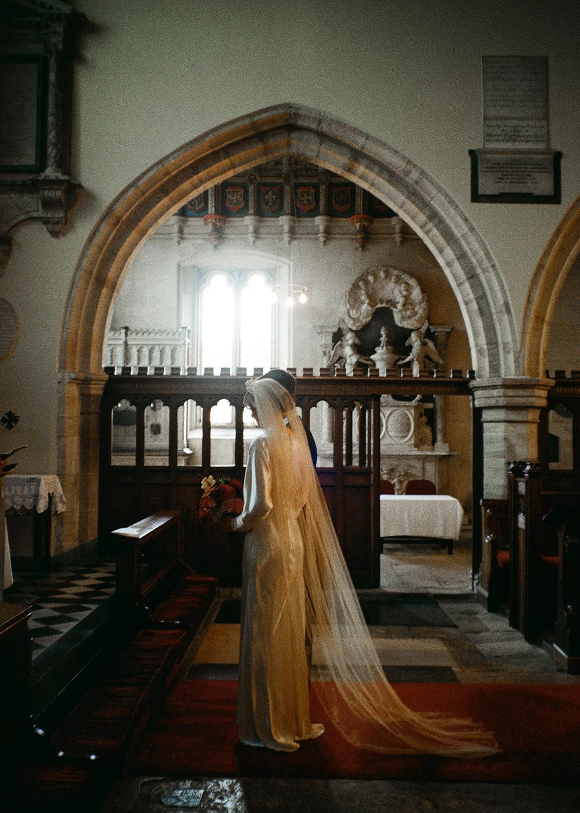 A bride in a long veil and cream wedding dress standing inside a church, holding a bouquet of flowers, facing a book on a stand, with a window in the background letting in light.