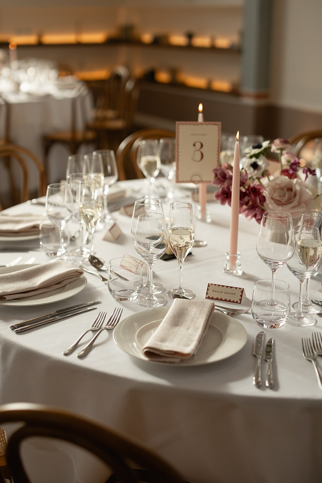 Elegant banquet table setup with white tablecloth, champagne glasses, water glasses, cutlery, napkins, pink floral centerpiece, candles, and table number 3, in a well-lit room.
