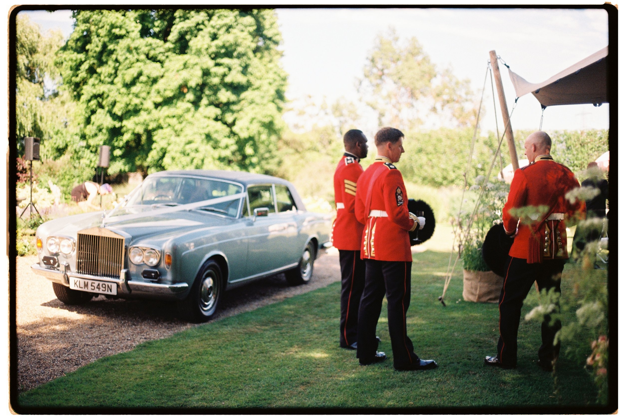 Men in red uniforms standing outdoors near a vintage car with a green garden and trees in the background.