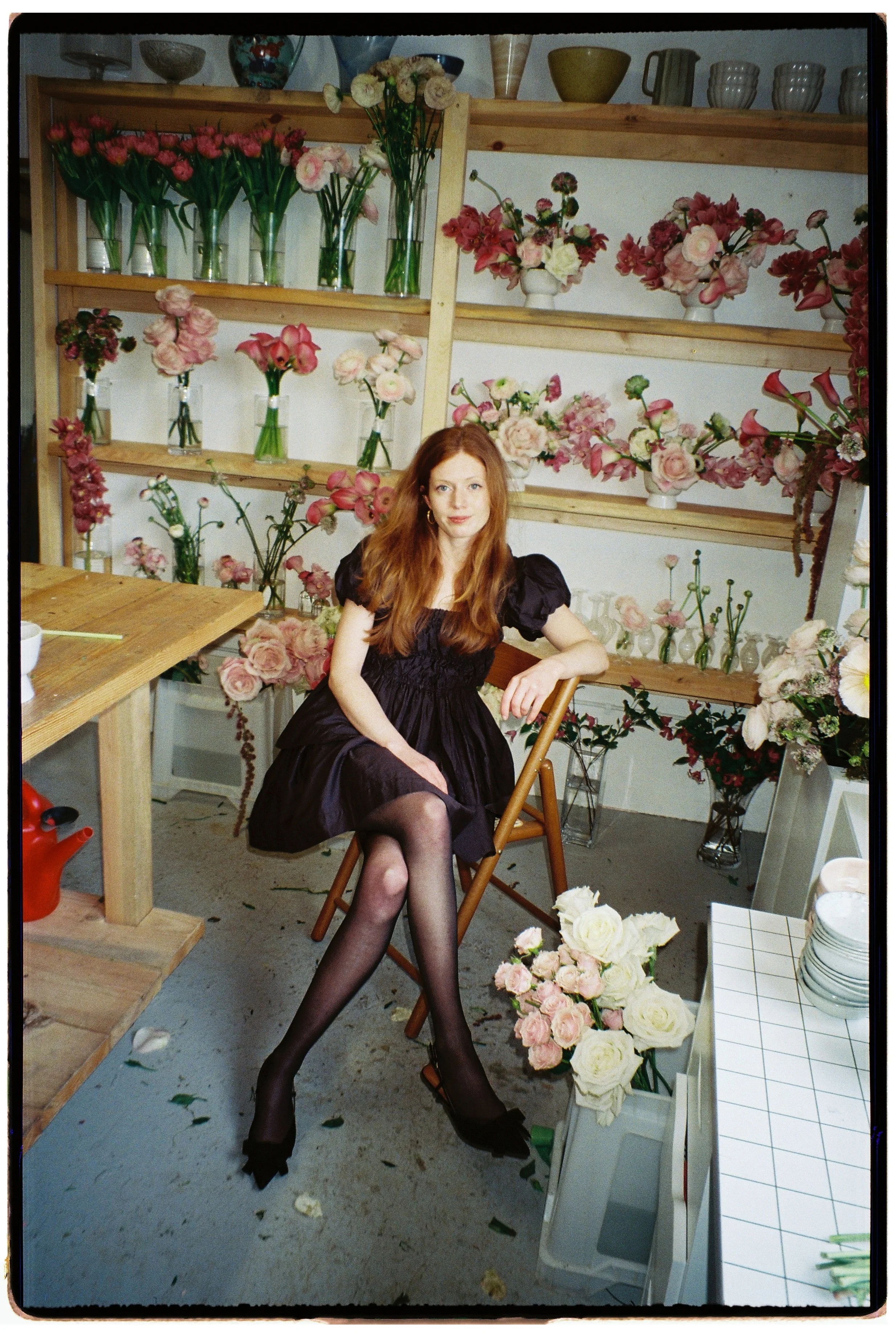 A woman with long auburn hair in a black dress and black stockings sits on a wooden chair in a floral shop surrounded by various pink and white flowers in vases and on shelves.